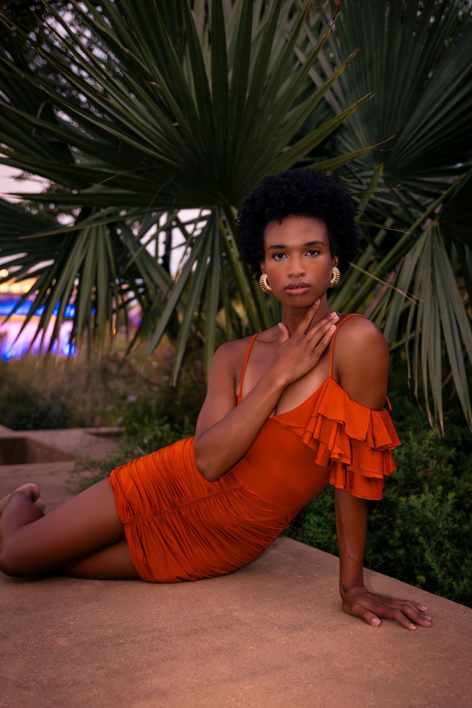 Woman in orange ruffled mini dress posing gracefully beside tropical palm trees at dusk.