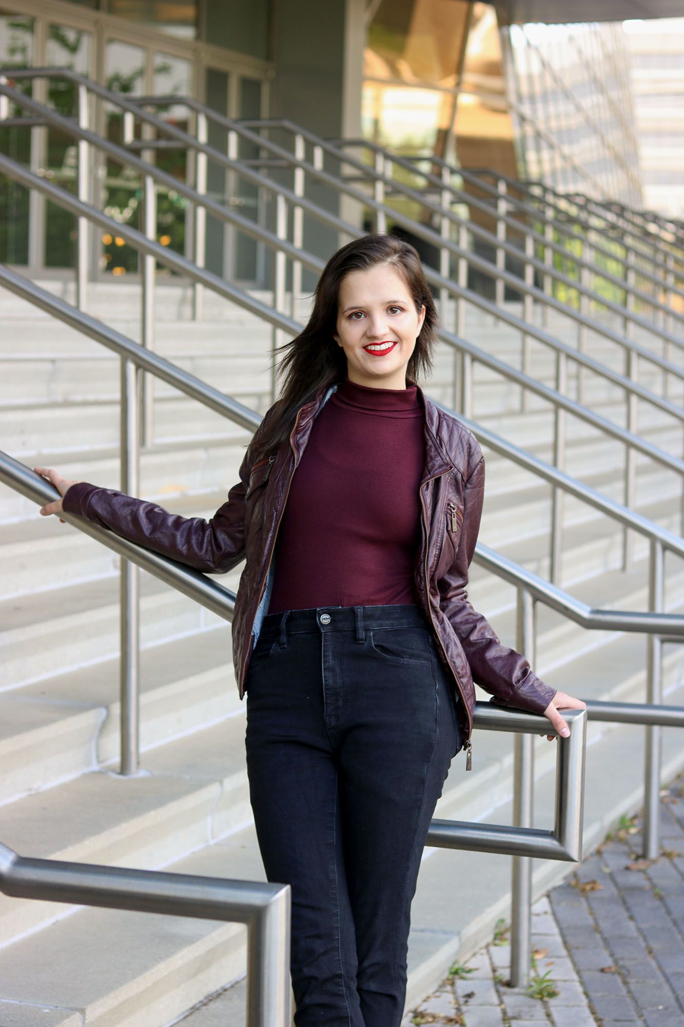Young woman in burgundy jacket and black jeans posing on outdoor staircase with metal railings.