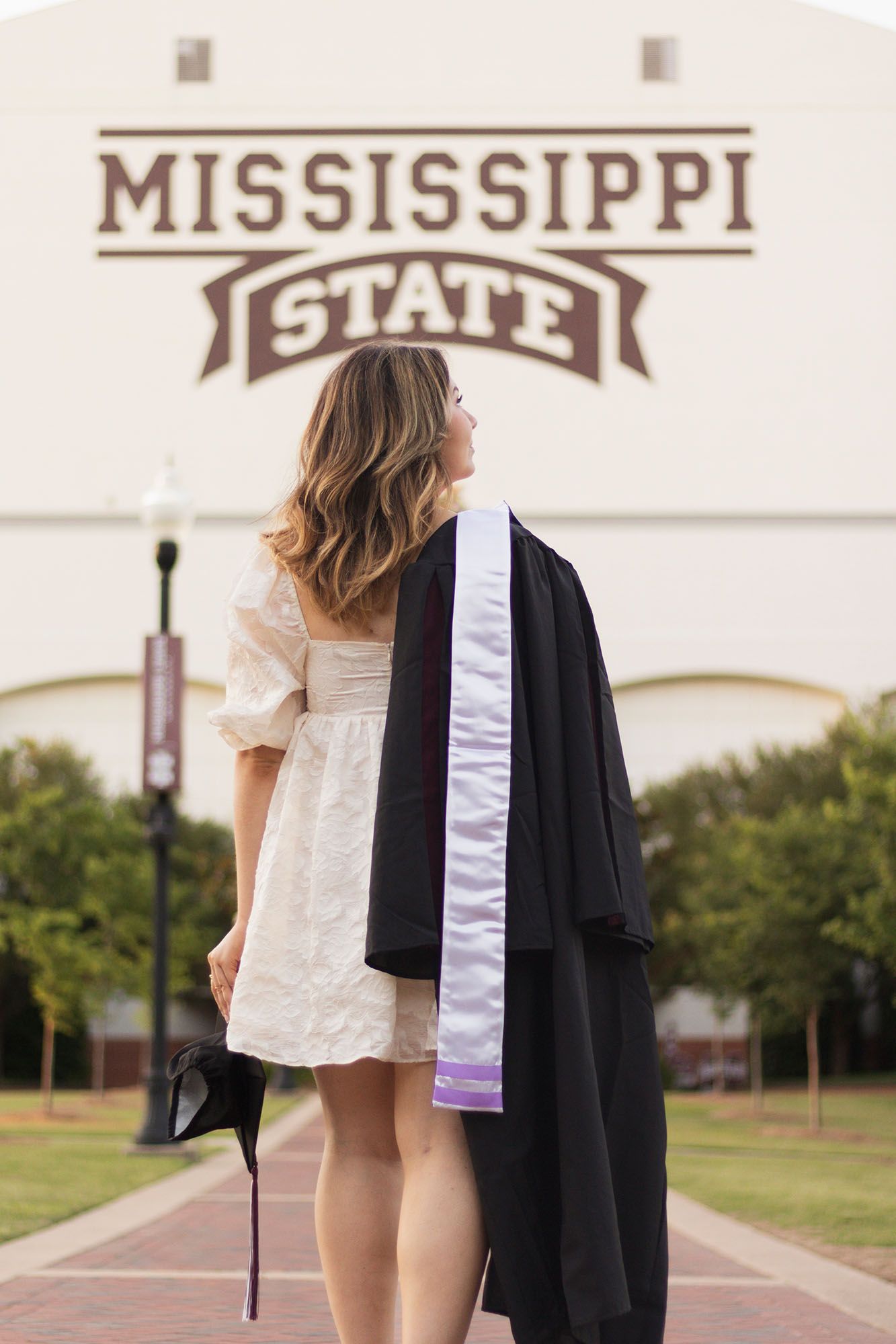 Graduate in white dress holds cap and gown facing Mississippi State university building.