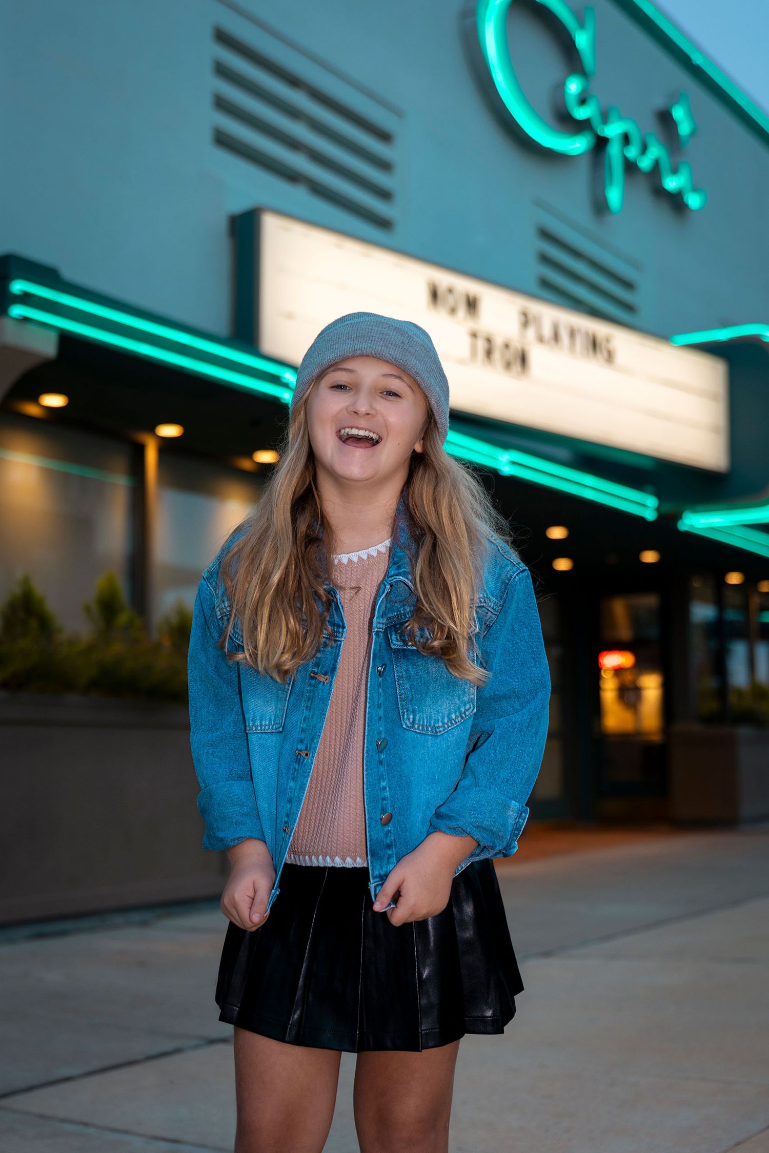 Smiling young girl in denim jacket standing outside the Capri Theatre at dusk.