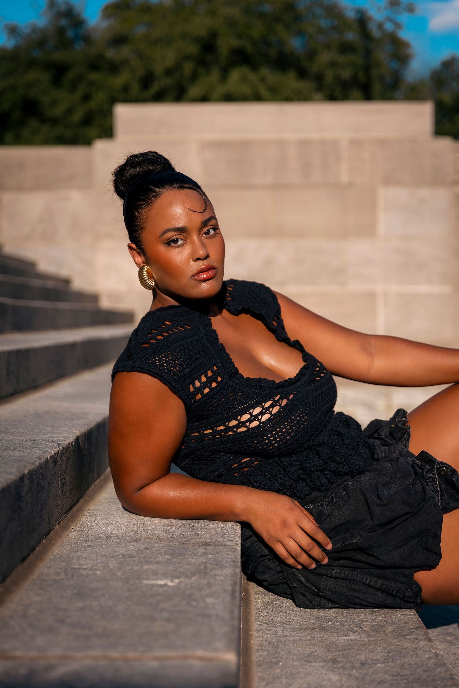 Woman in black crochet top and skirt poses on stone steps in sunlight.