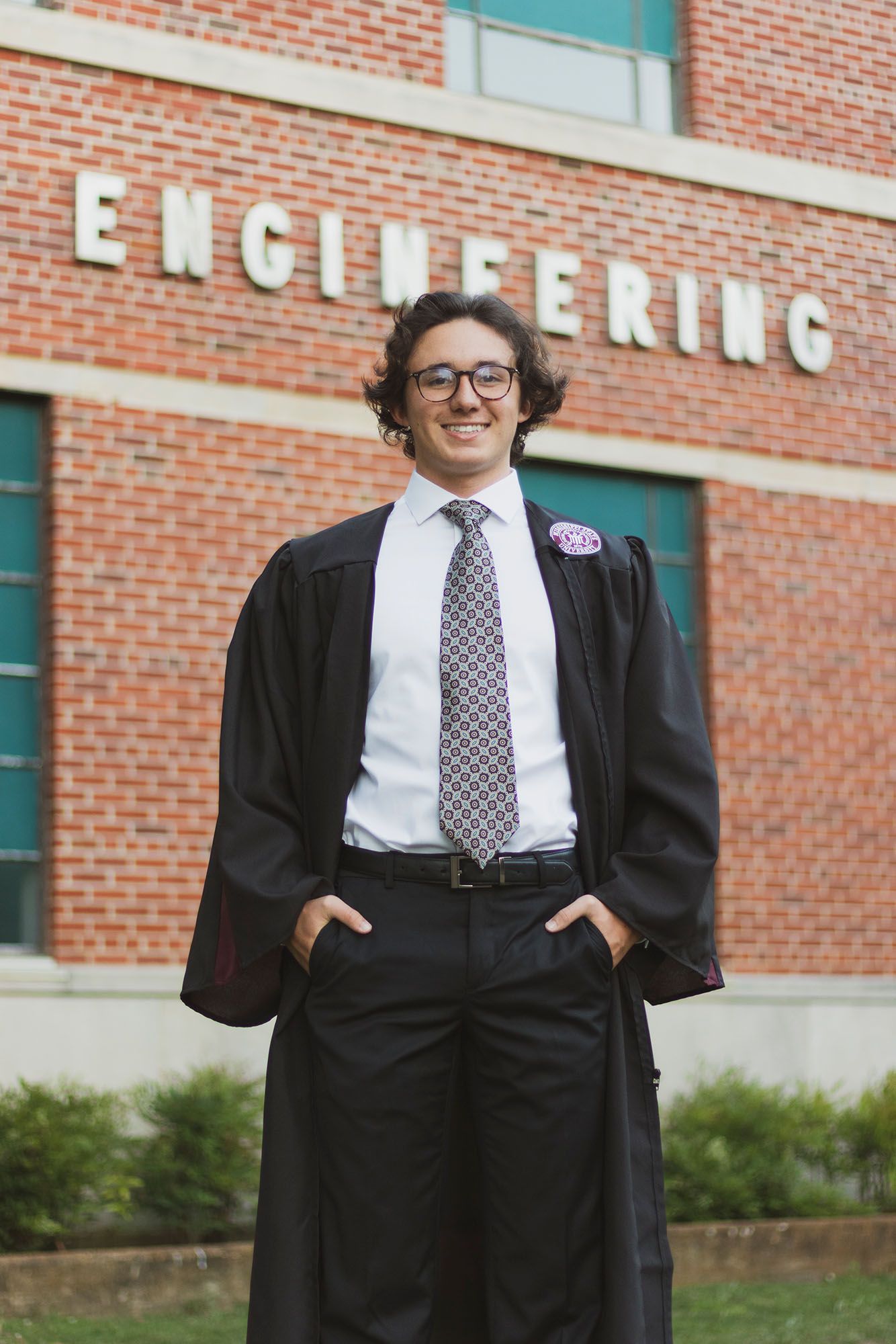 Smiling graduate in black graduation gown standing in front of brick Engineering building on campus.