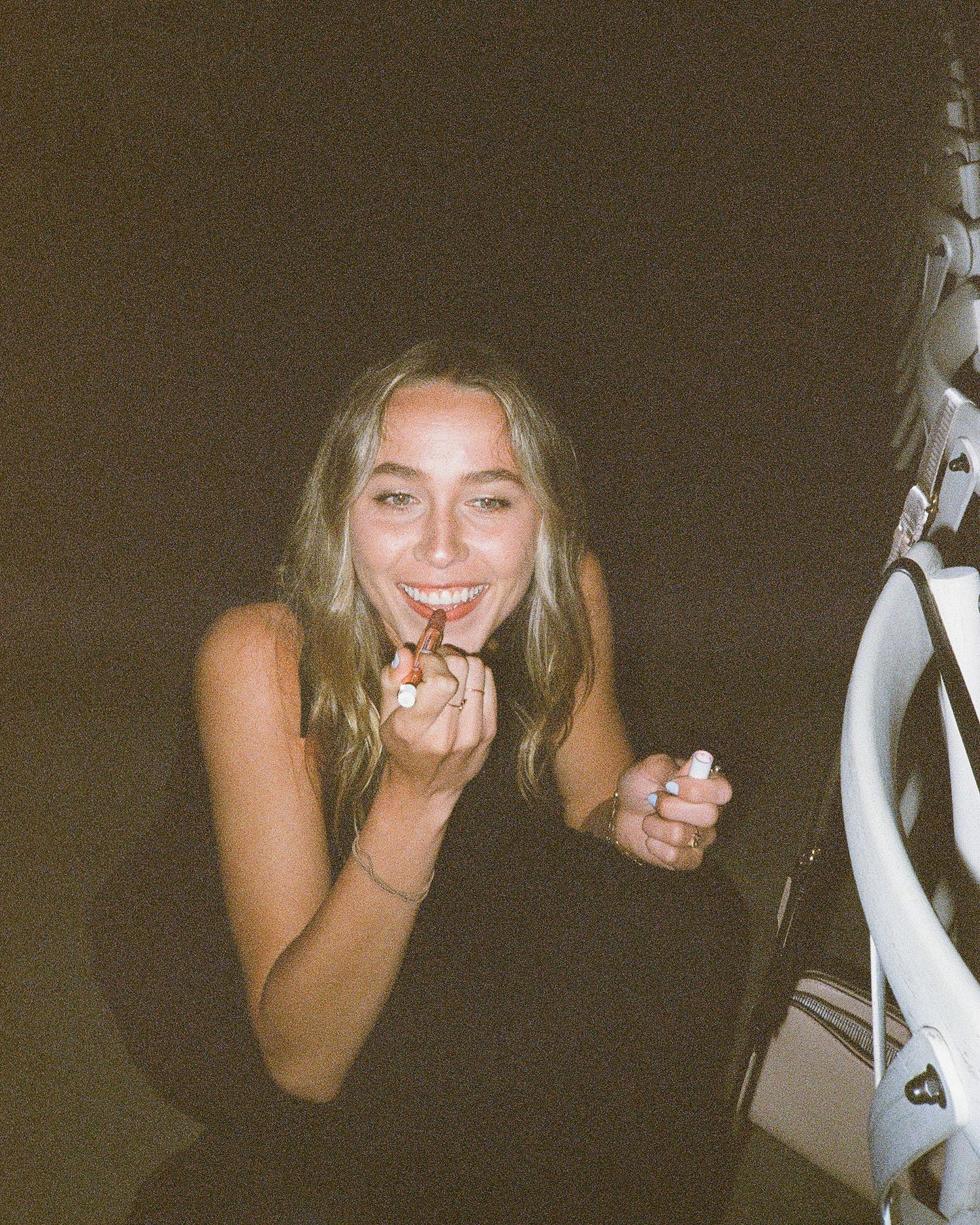 Young blonde woman applying lipstick and smiling in a dark setting.