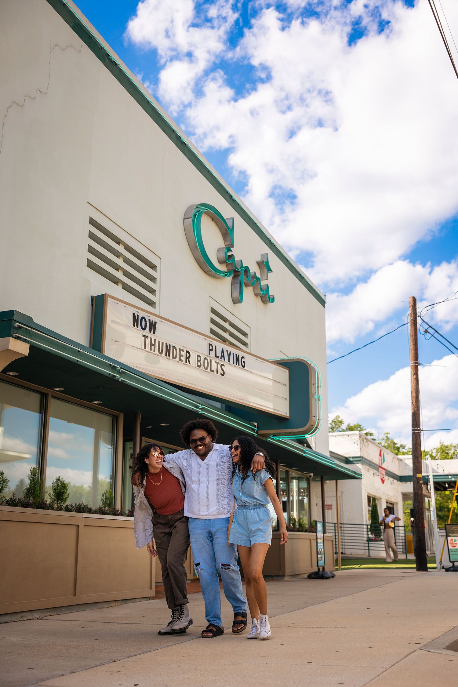 Three friends laugh and walk together outside the Capri Theater showing Thunder Bolts on a sunny day.