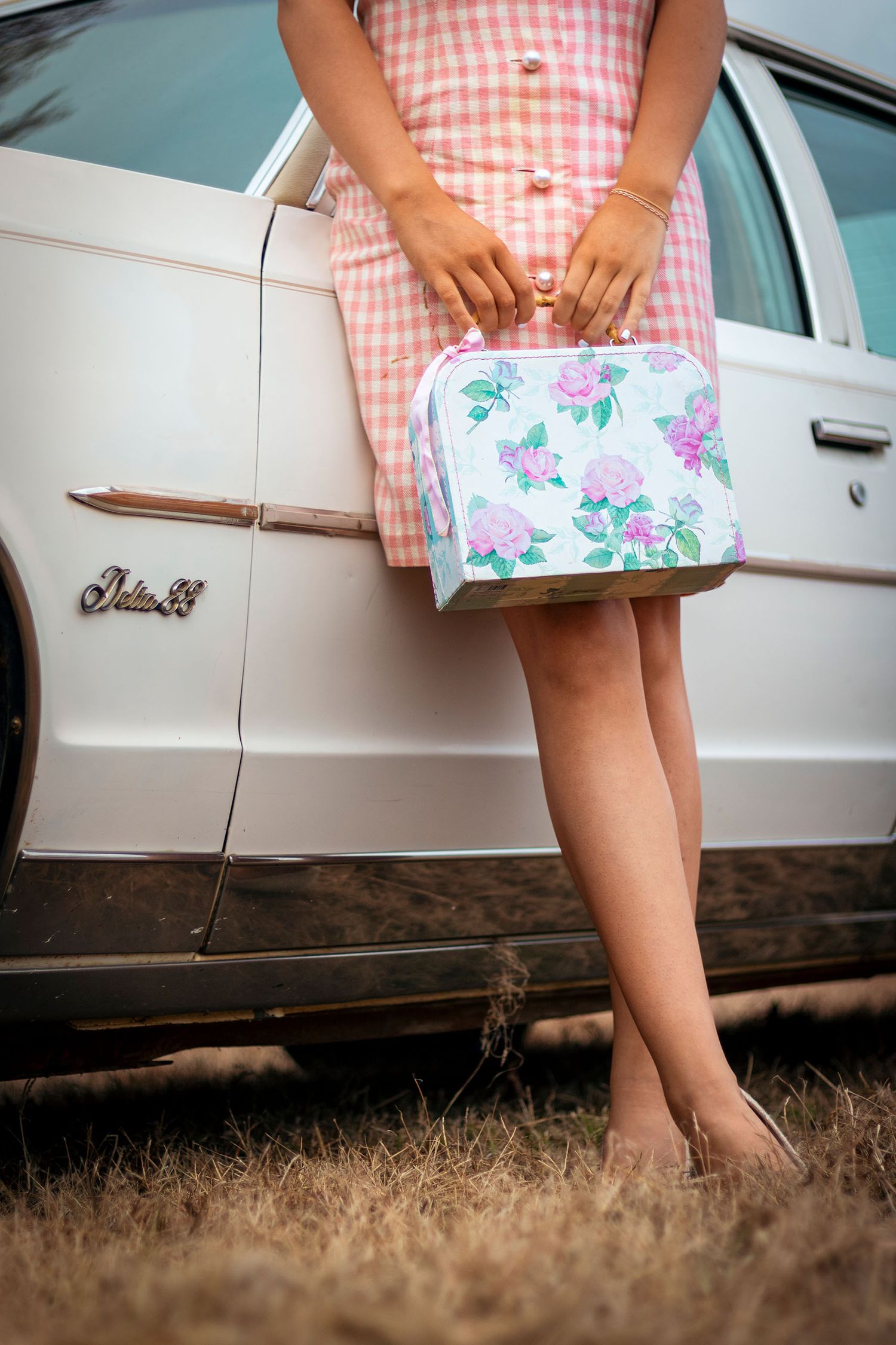 Woman in pink gingham dress holds floral cosmetic bag beside vintage white Oldsmobile car.