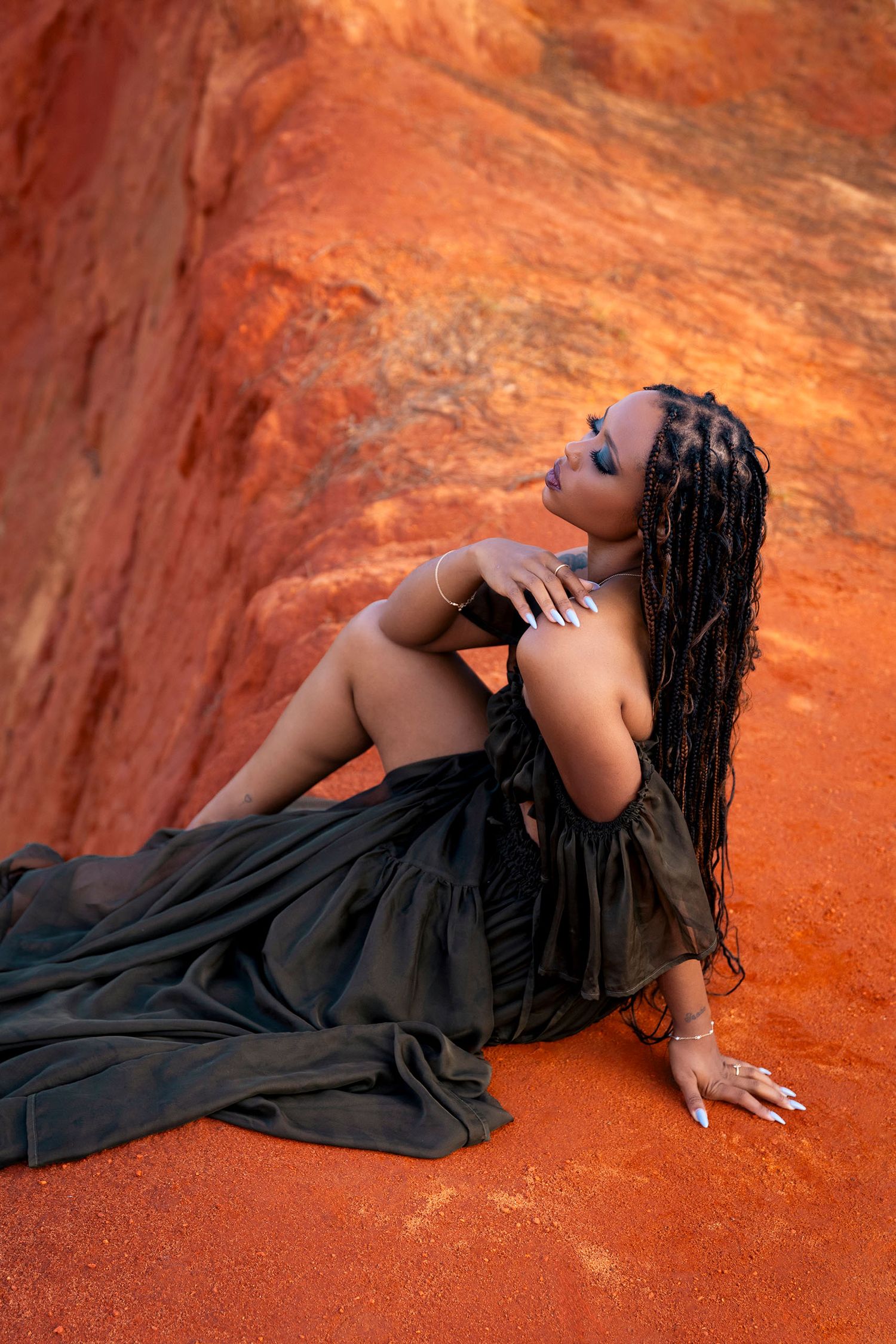 Woman in black dress poses against red rock desert backdrop with long braided hair.