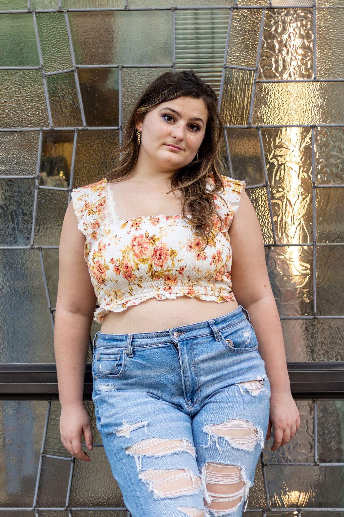 Young woman in floral crop top and ripped jeans posing against a stained glass wall.