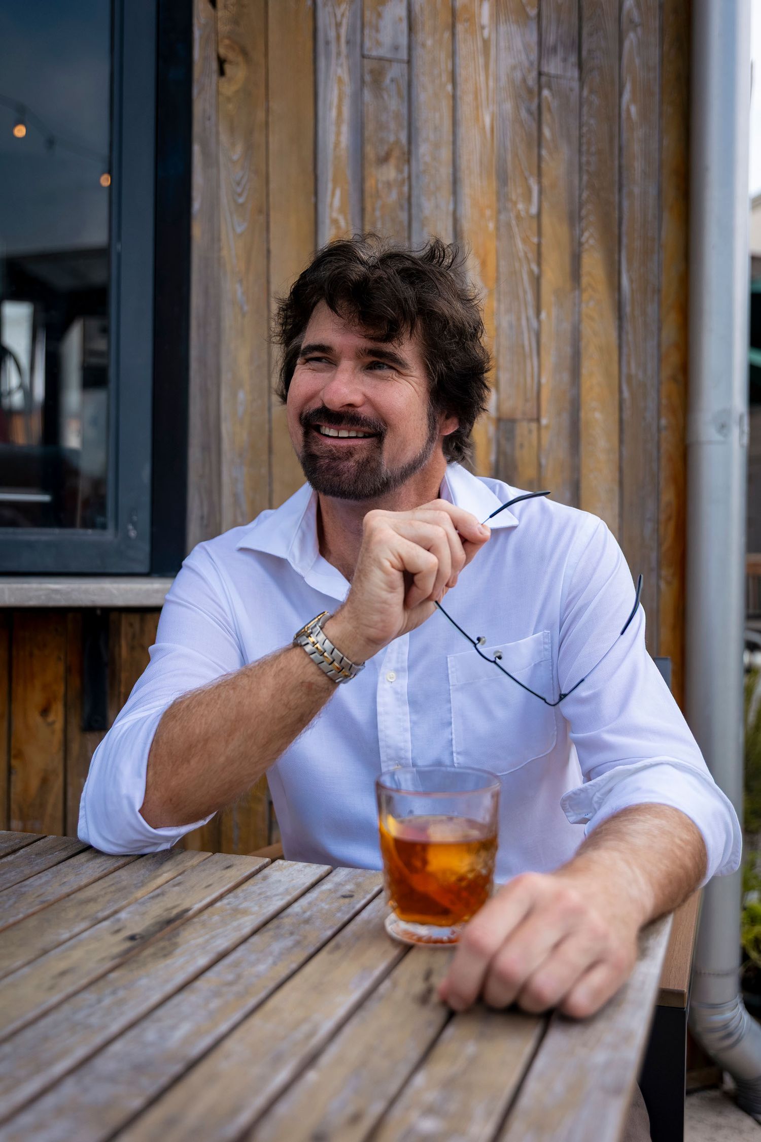 A smiling bearded man in a white shirt sits at an outdoor wooden table with an iced drink.