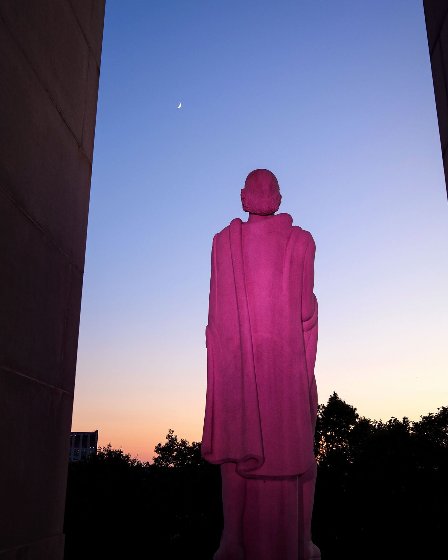 Pink illuminated statue silhouetted against a twilight sky with a crescent moon.