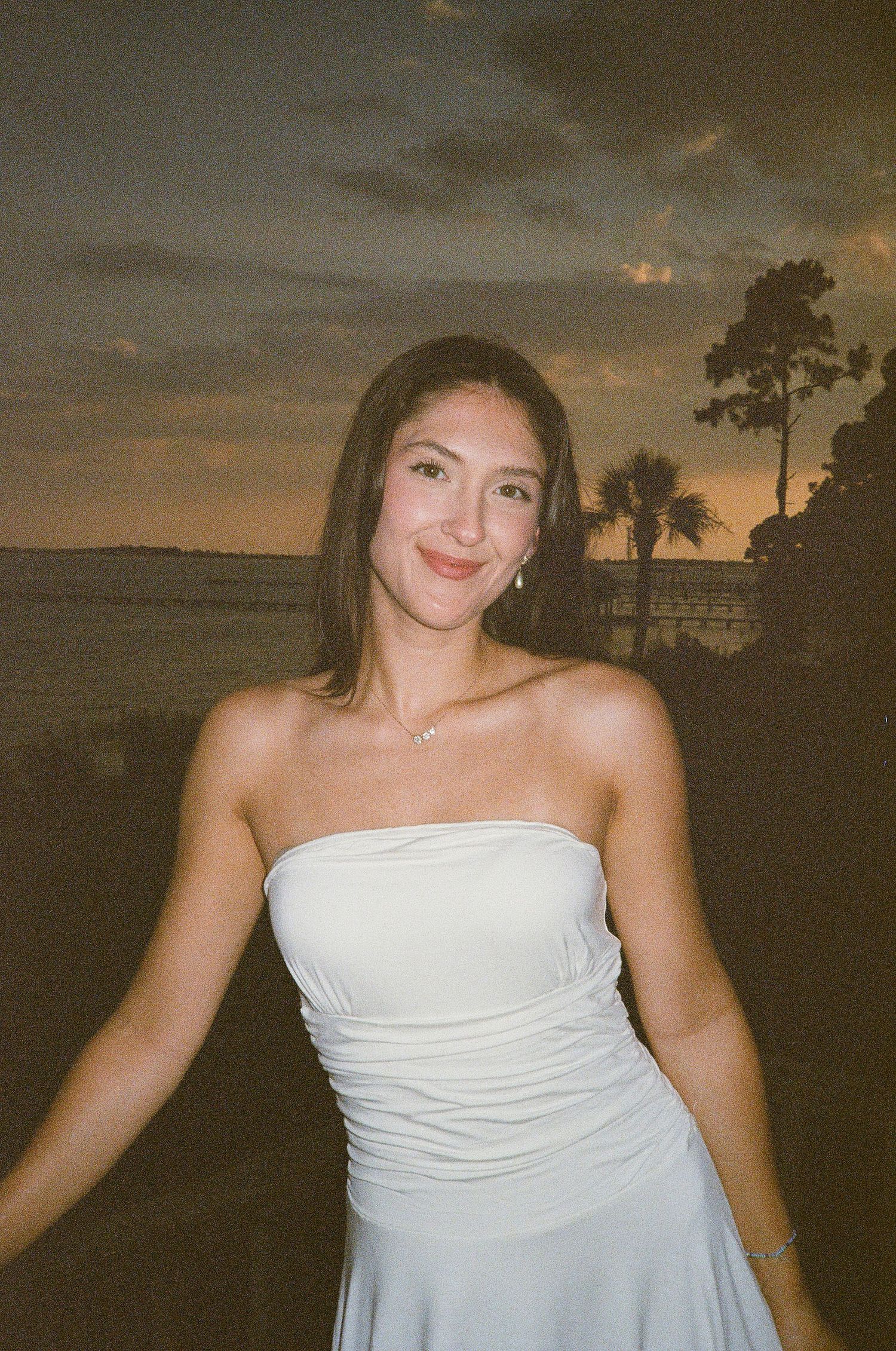 Young woman in a white strapless dress smiling at sunset near a waterfront with palm trees.