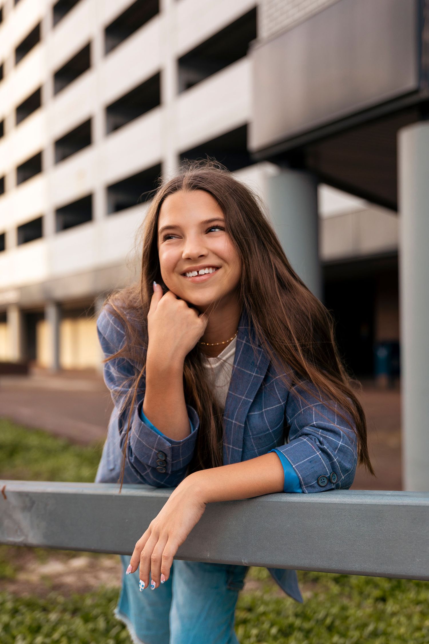 Smiling young woman with long brown hair wearing a blue plaid blazer, leaning on a railing outdoors.