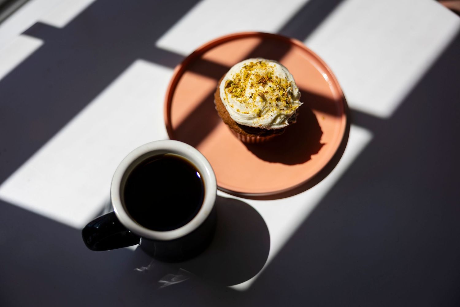 A black coffee mug beside a frosted cupcake on an orange plate in dramatic natural sunlight.