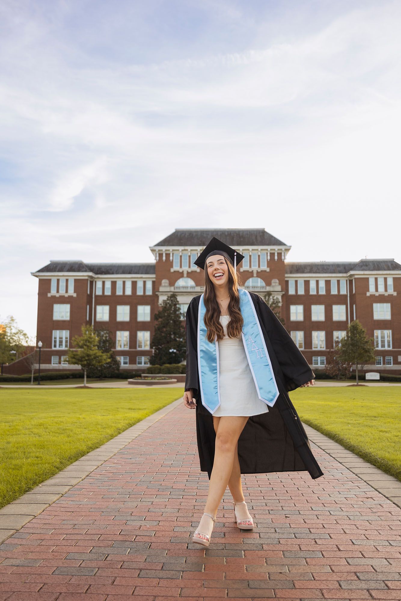 Joyful graduate in cap and gown walking on brick path in front of brick university building.