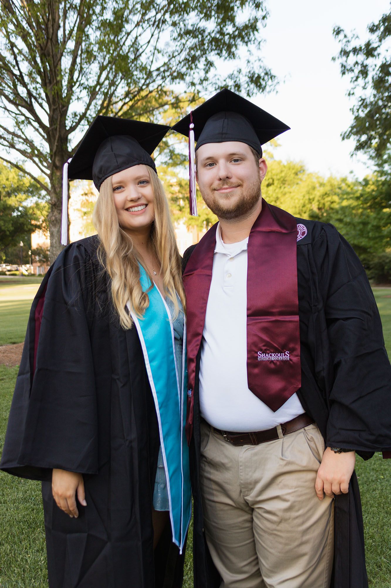 Two smiling graduates in black caps and gowns pose together on green campus lawn at dusk.