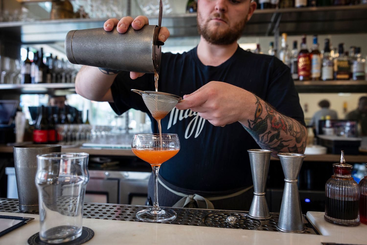 Tattooed bartender strains an orange cocktail into a glass at a well-stocked bar.
