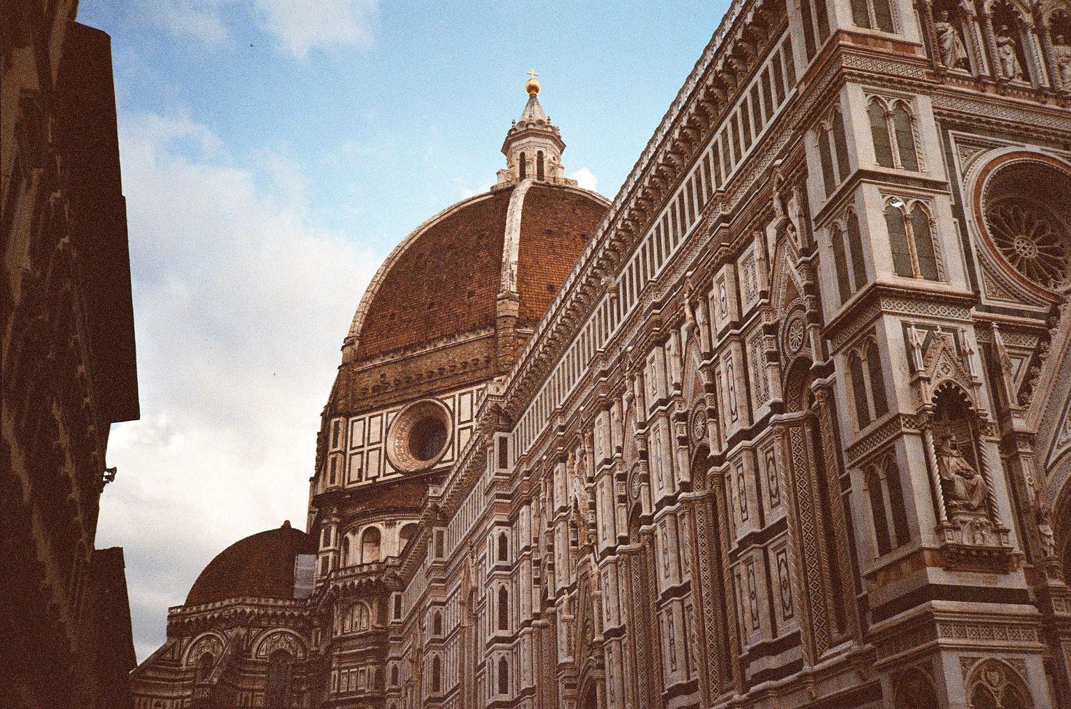 The Florence Cathedral dome and ornate marble facade rise against a partly cloudy sky in Italy.