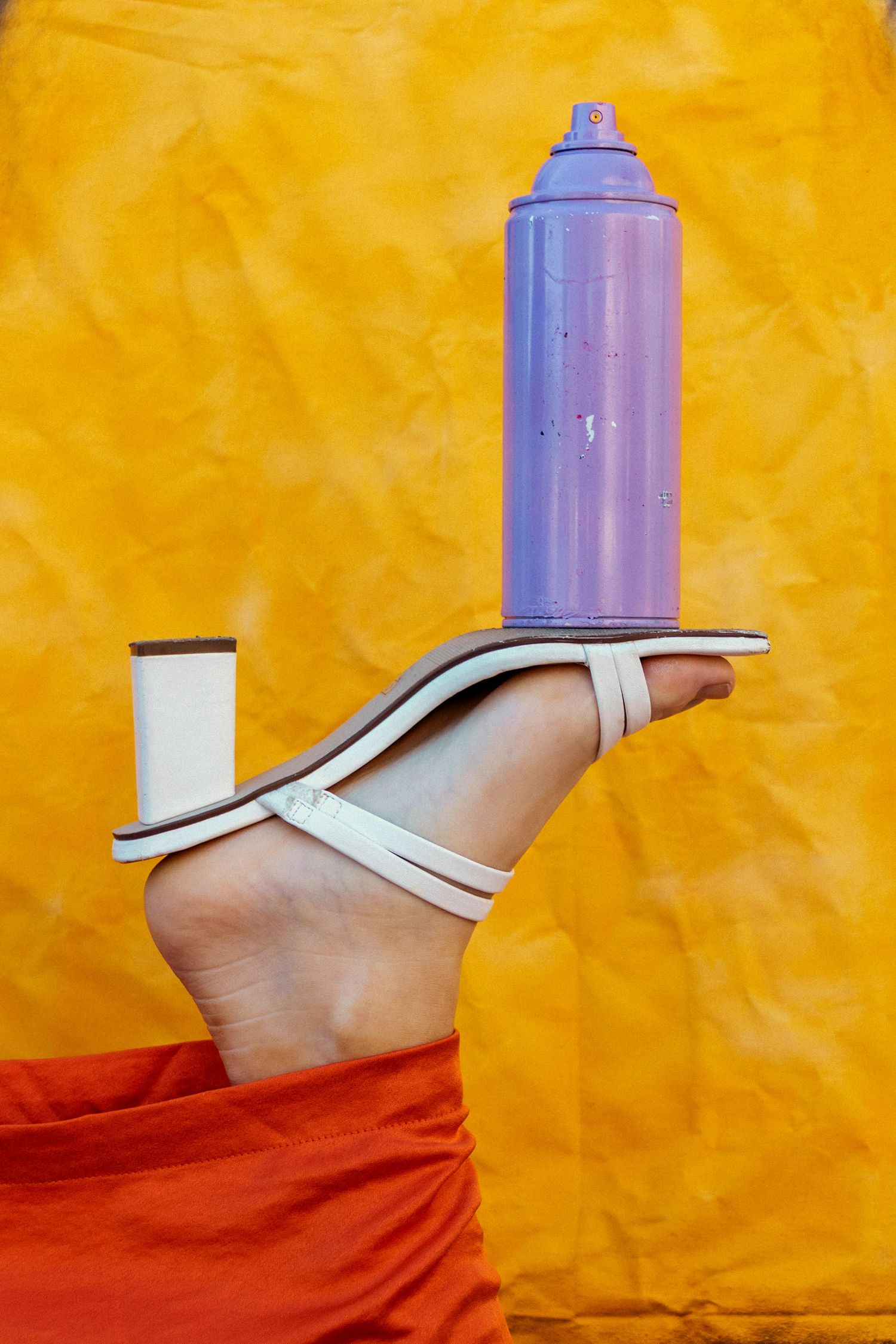 Foot wearing white strappy heeled sandal balancing purple spray can against bright yellow background.