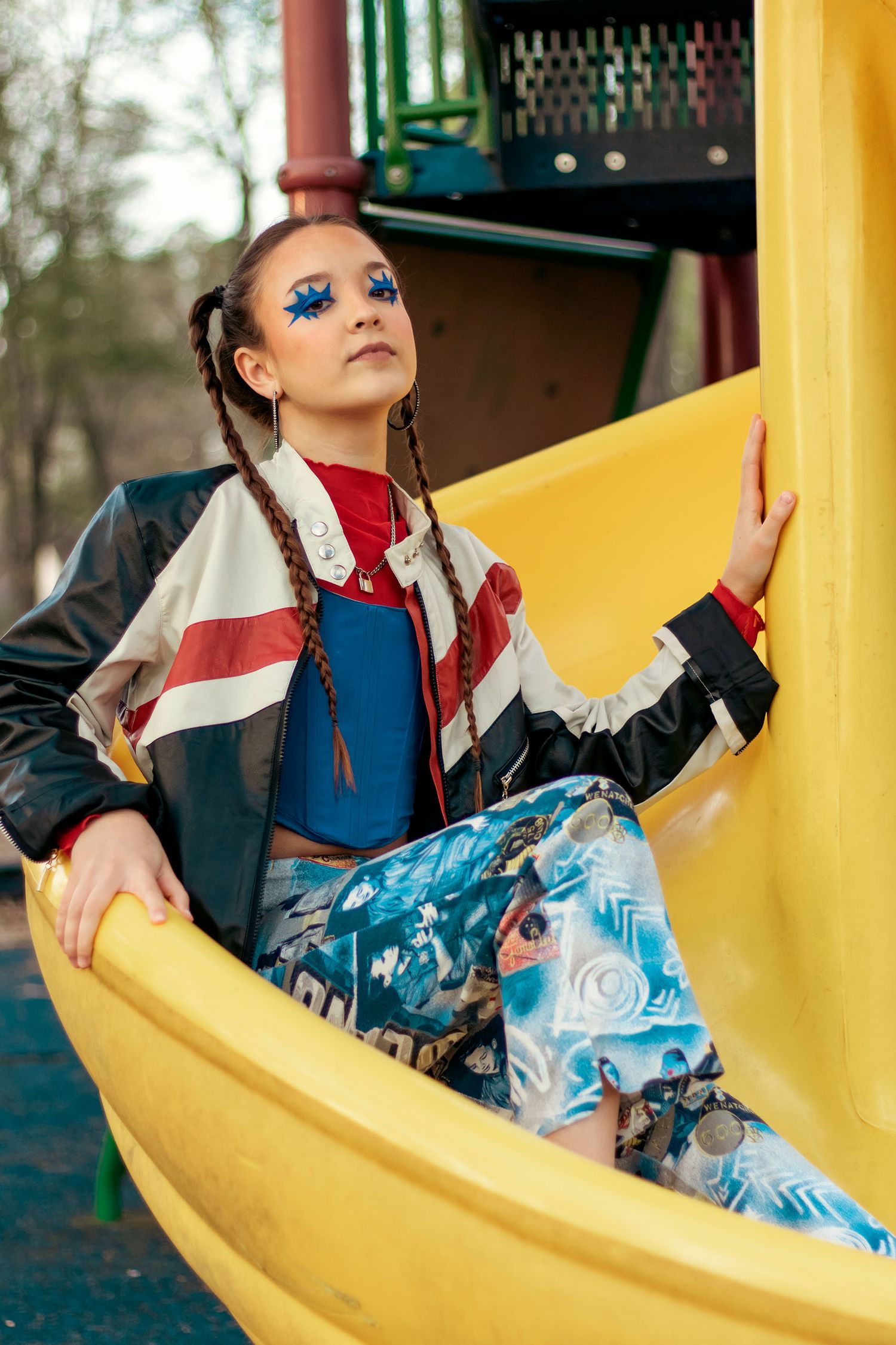 Young girl in colorful streetwear with blue glitter eye makeup posing on a yellow playground slide.