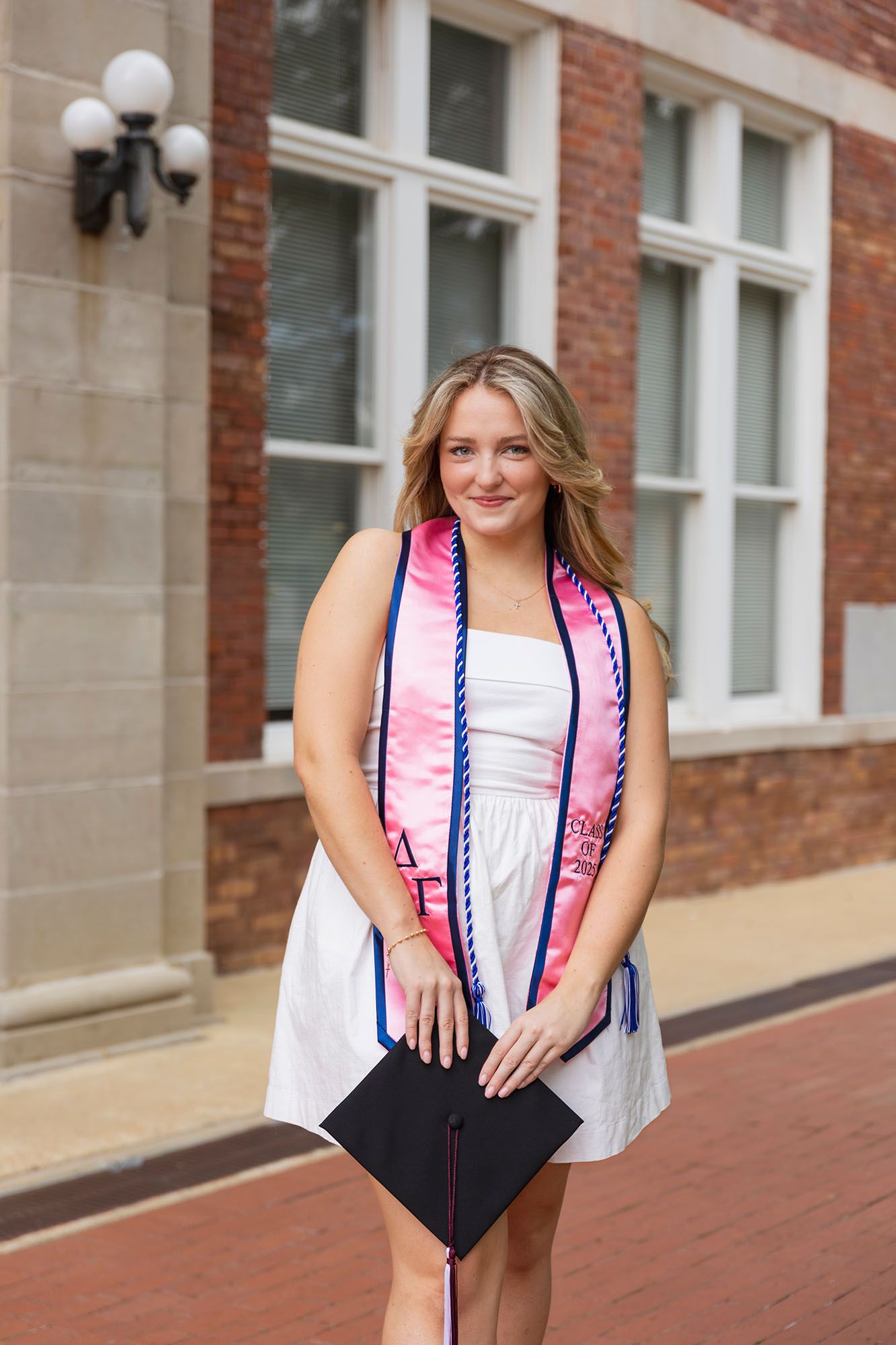 Smiling female graduate in white dress with pink sorority stole holds cap at brick building.