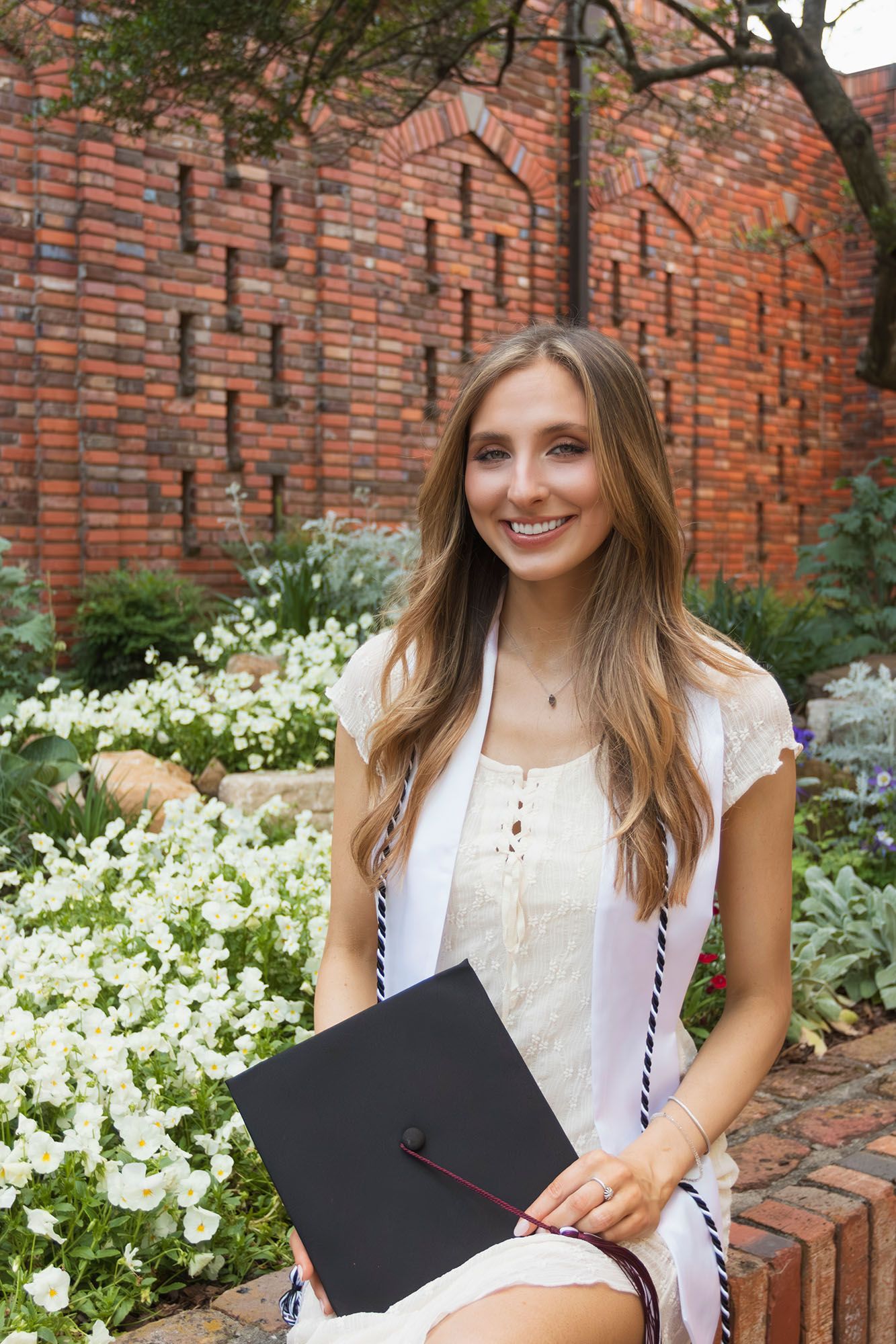 Smiling female graduate holds cap while seated among white flowers at brick university garden.