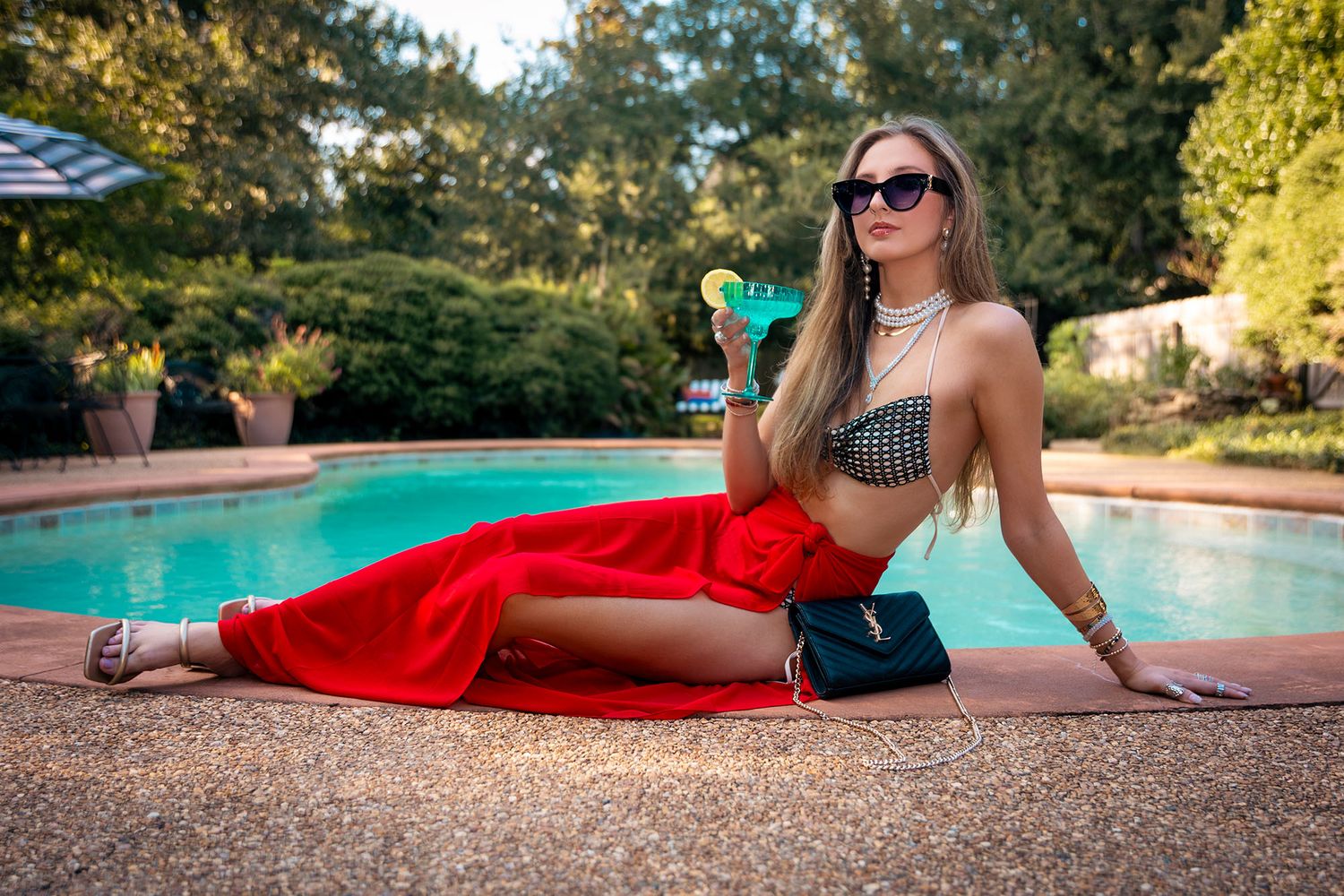 Blonde woman in red maxi skirt and printed bikini top sitting poolside wearing sunglasses while drinking a beverage.