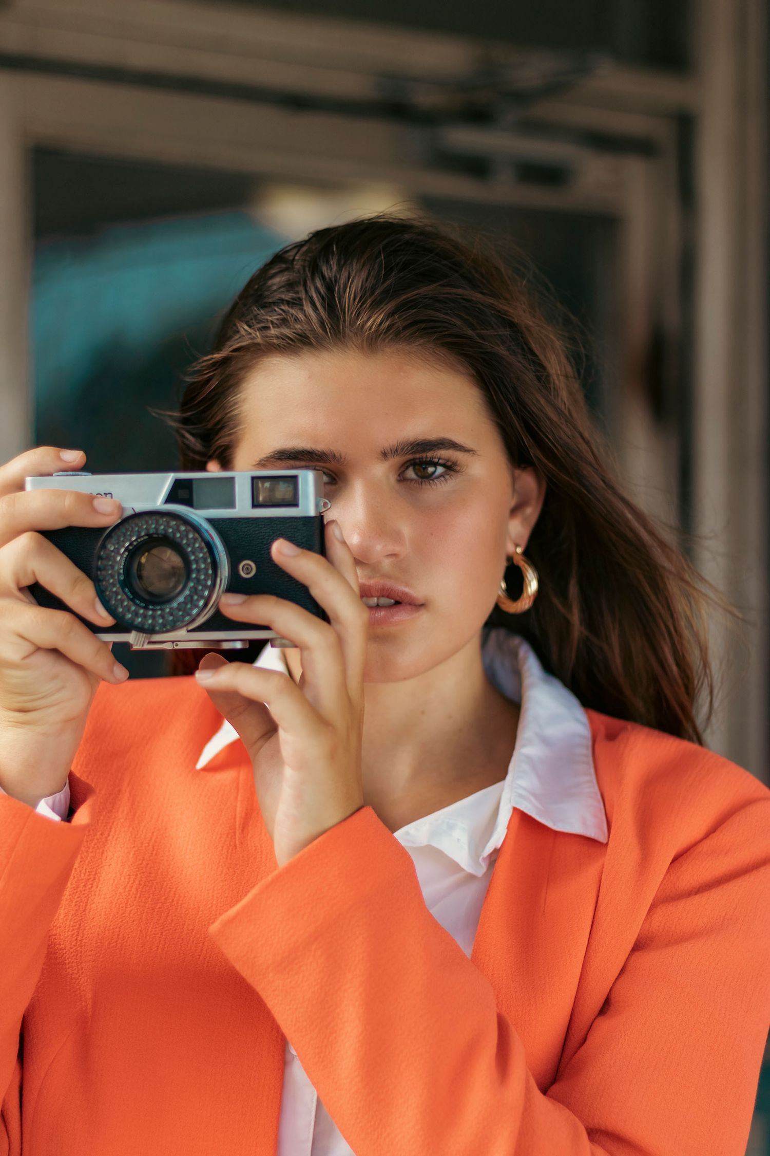 Young woman in orange blazer holds a vintage film camera up to her face outdoors.
