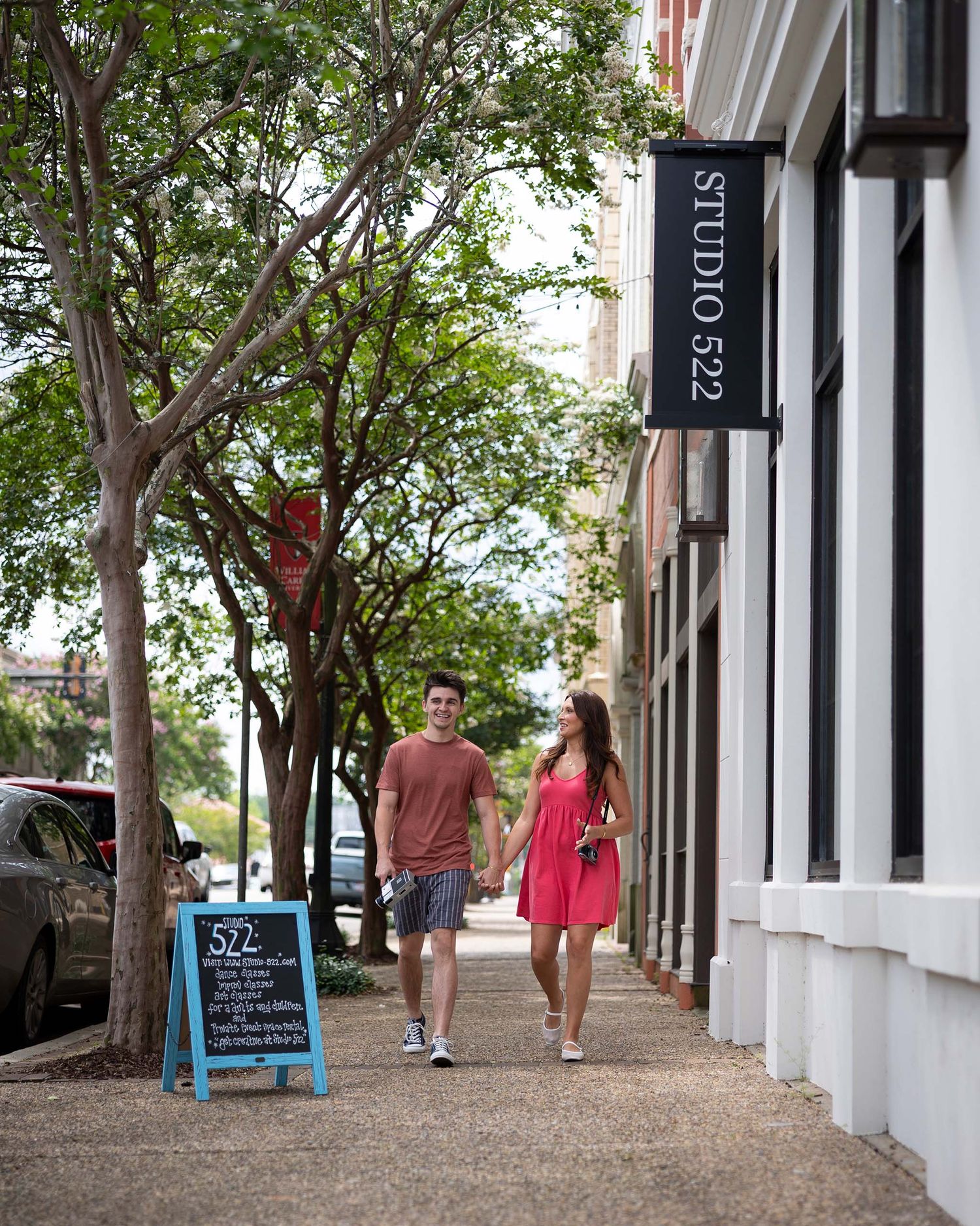 Couple holding hands walking past Studio 522 on a sunny tree-lined sidewalk.