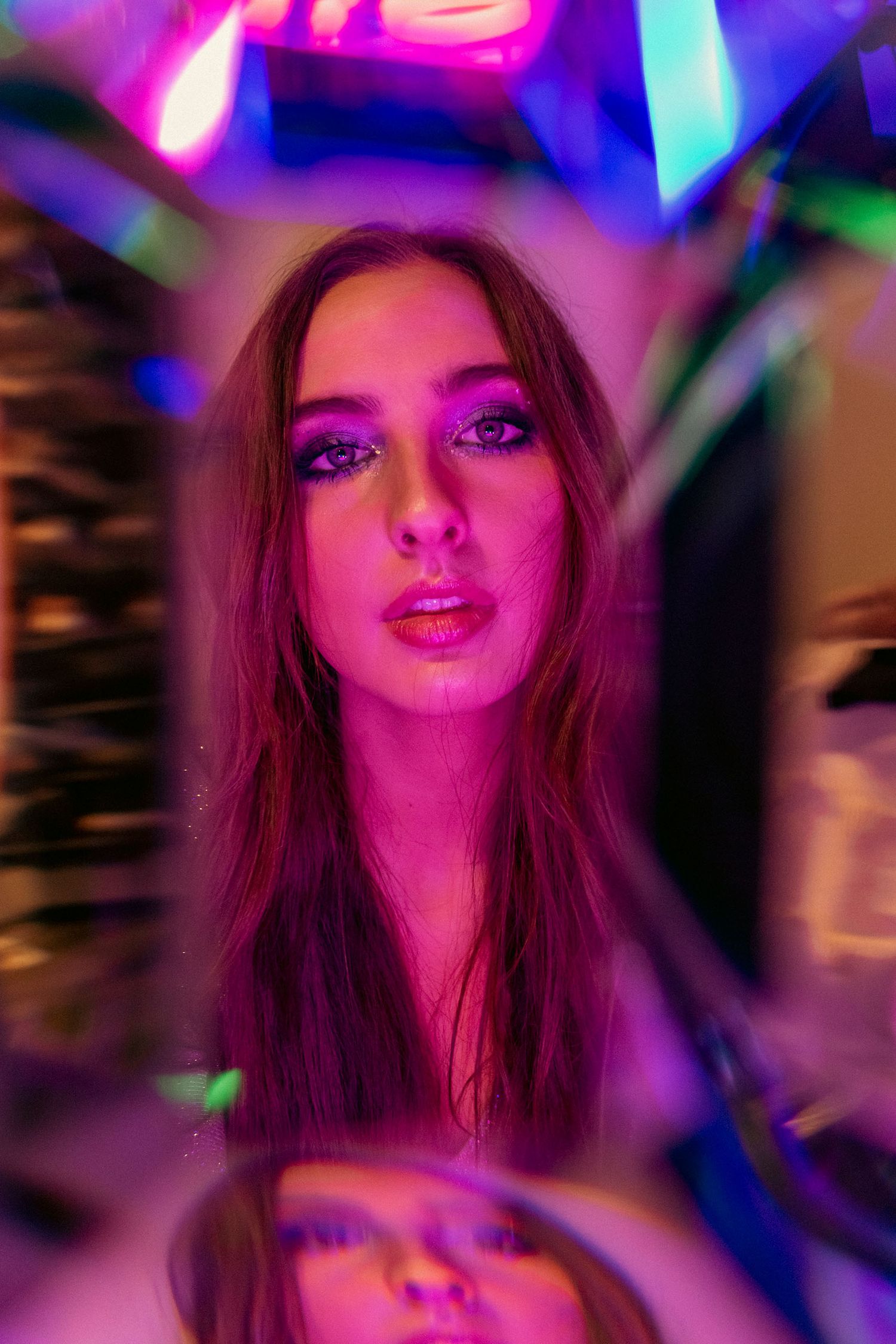 Young woman with auburn hair and pink makeup photographed through reflective material in neon-lit setting.