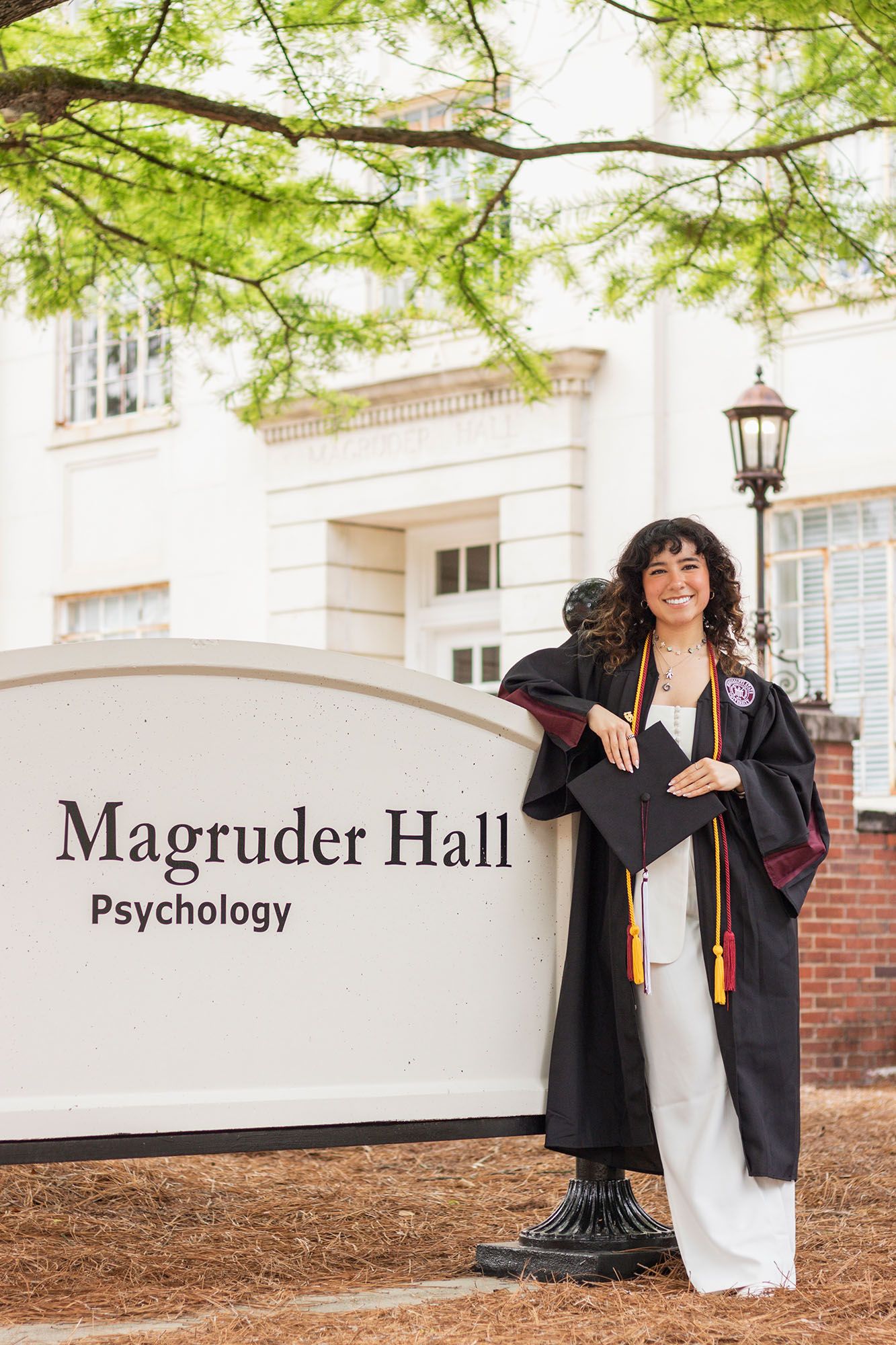Graduate in cap and gown poses beside Magruder Hall Psychology department sign at MSU.