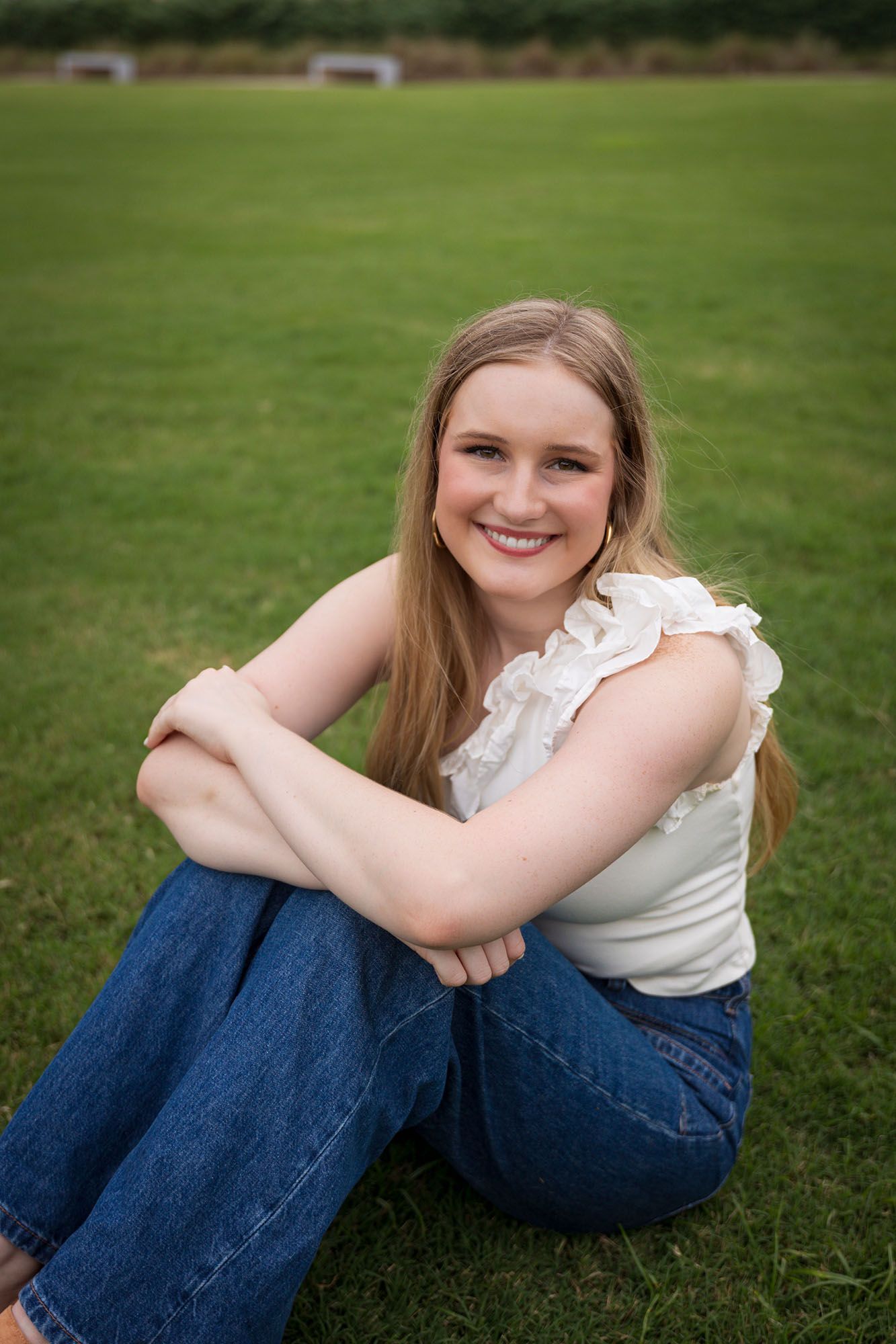 Smiling young woman in white ruffled top and jeans sits cross-legged on a green grass field.