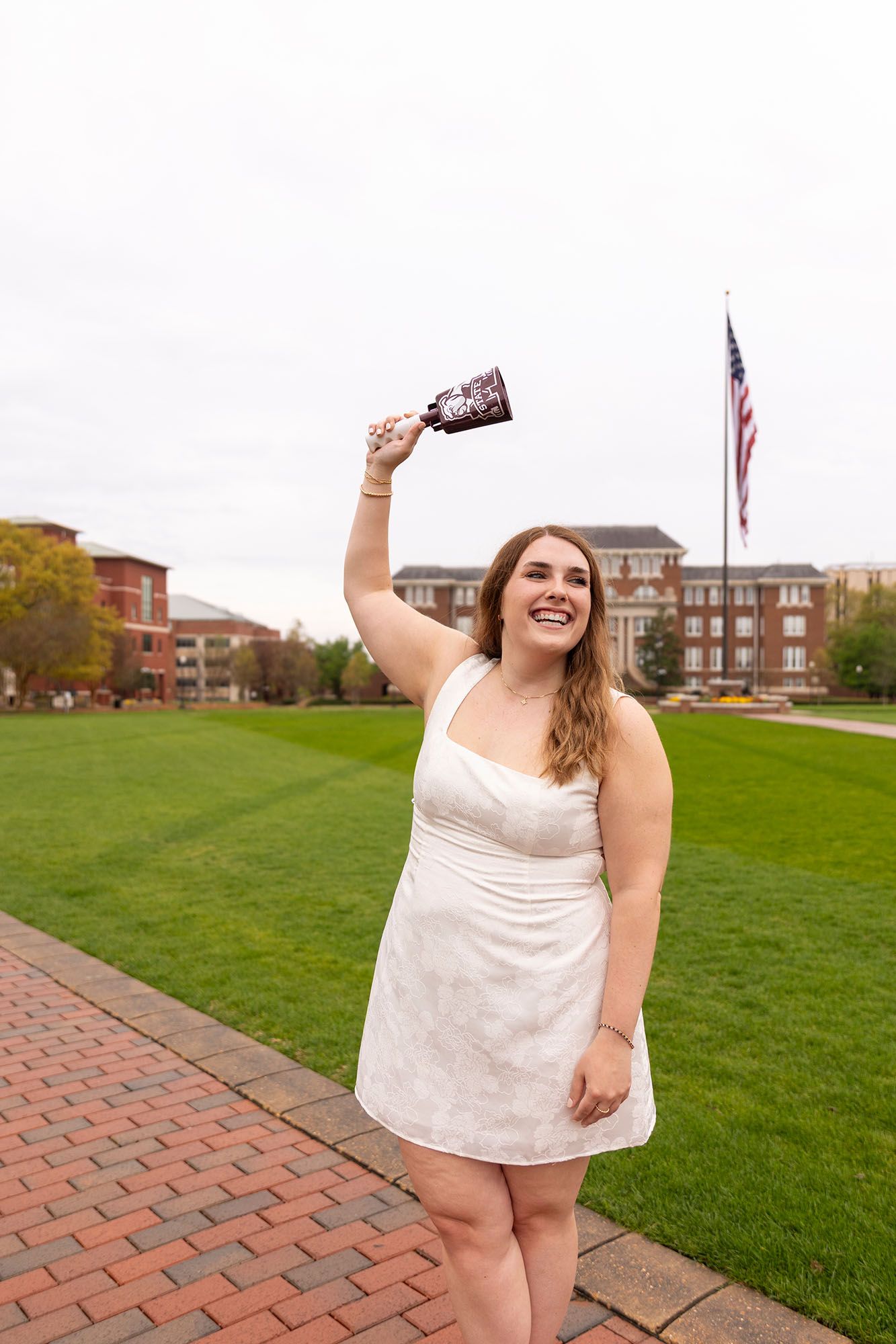 Happy graduate in white dress raising a cup on a brick campus path with American flag.