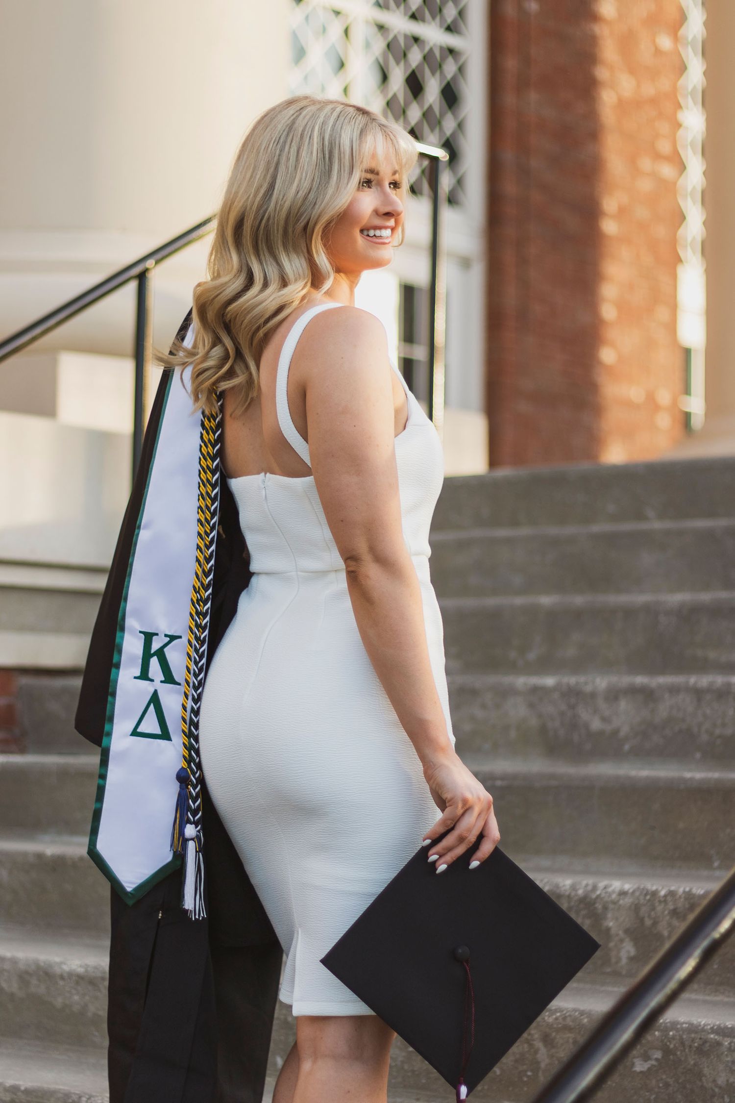 Blonde graduate smiles over shoulder holding cap and Kappa Delta stole on sunlit brick steps.