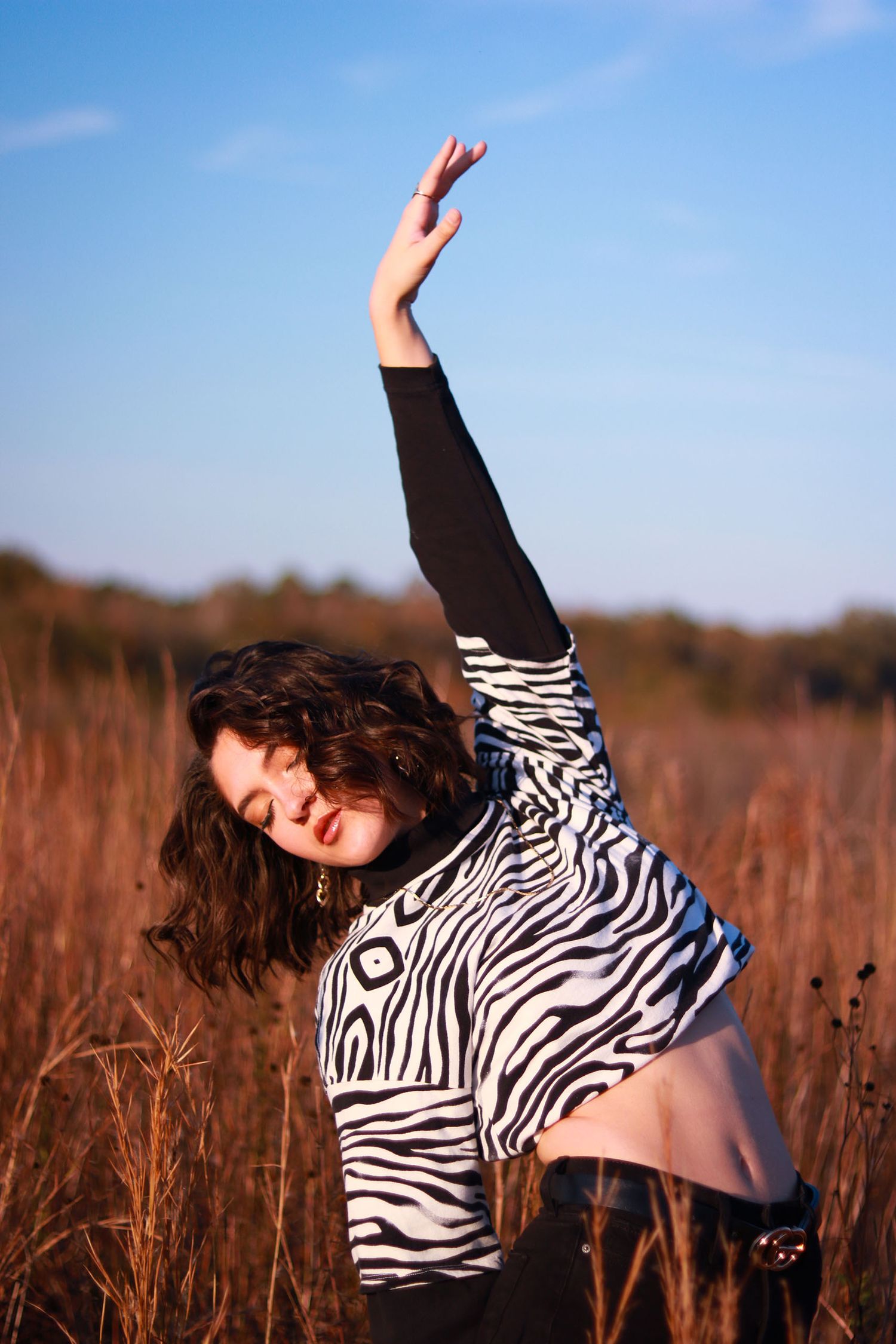 Woman in zebra print top raises arm dramatically in a golden field under blue sky.