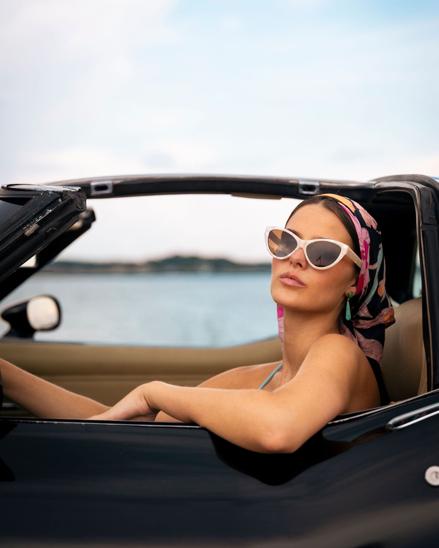 Stylish woman wearing white cat-eye sunglasses and floral headscarf sitting in black convertible.