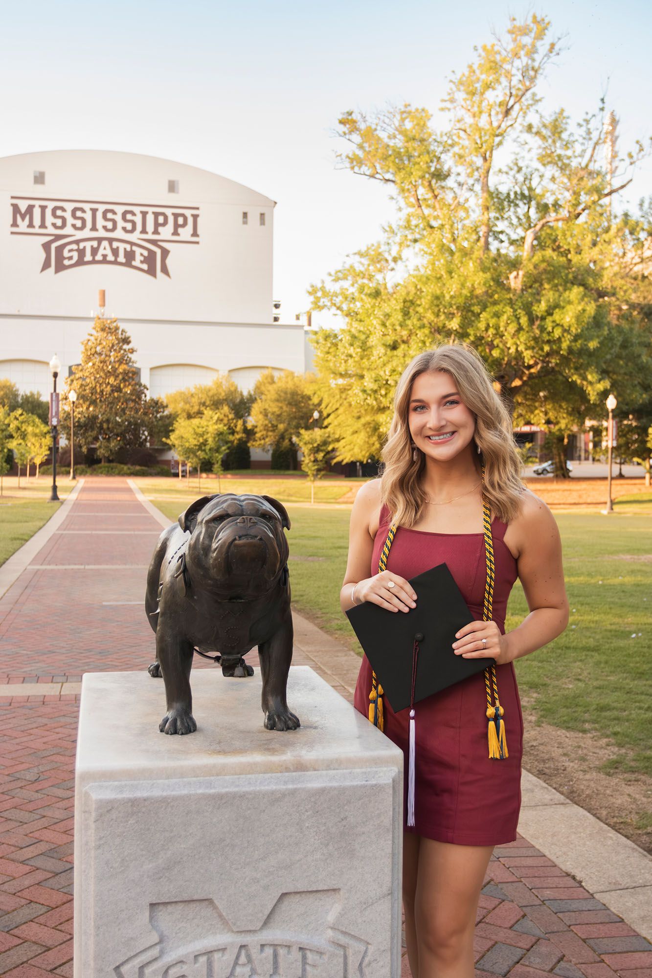 Female graduate poses with Mississippi State bulldog statue holding cap and honor cords.