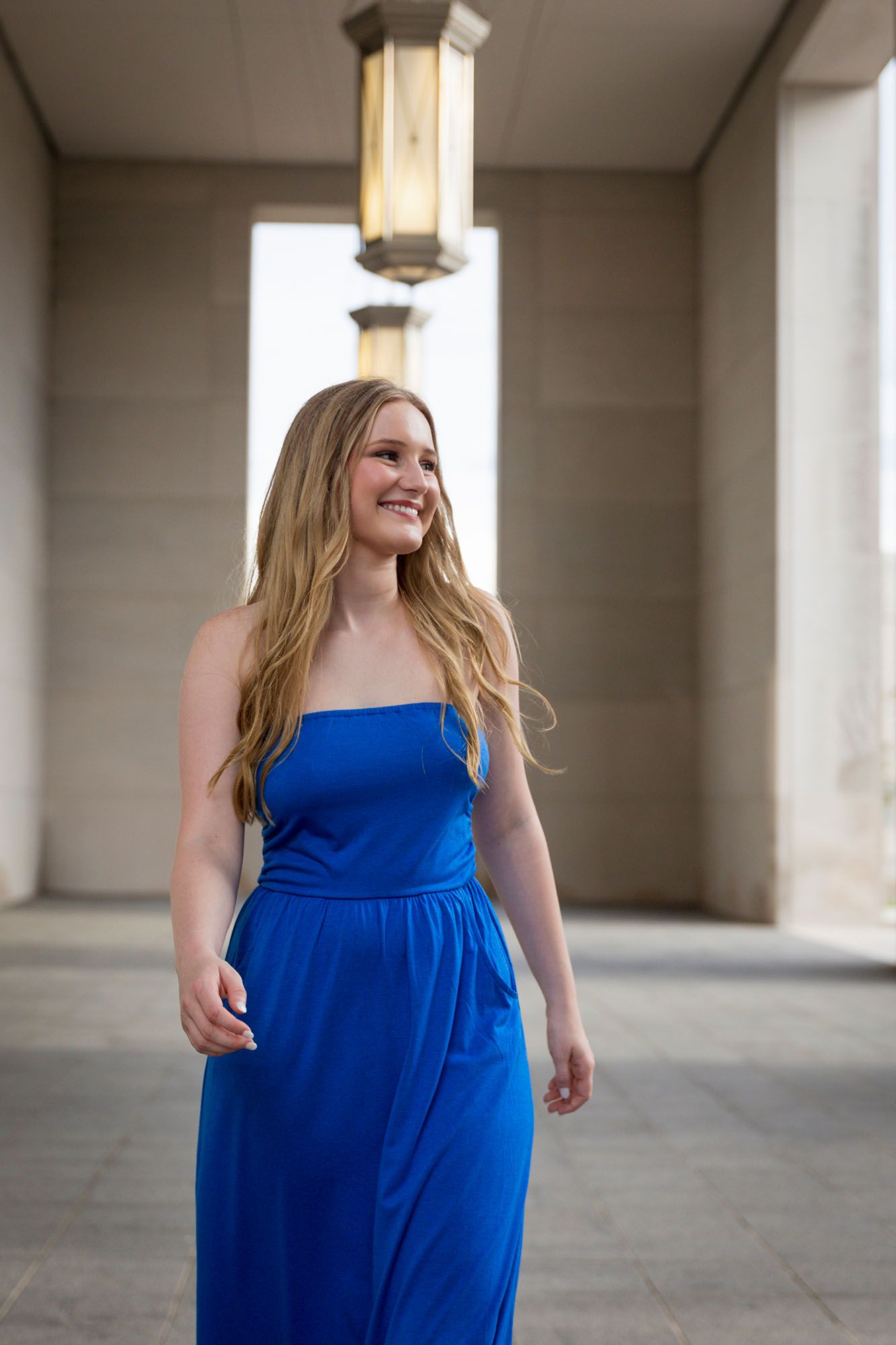 Young blonde woman in a strapless royal blue maxi dress walks through an elegant corridor.