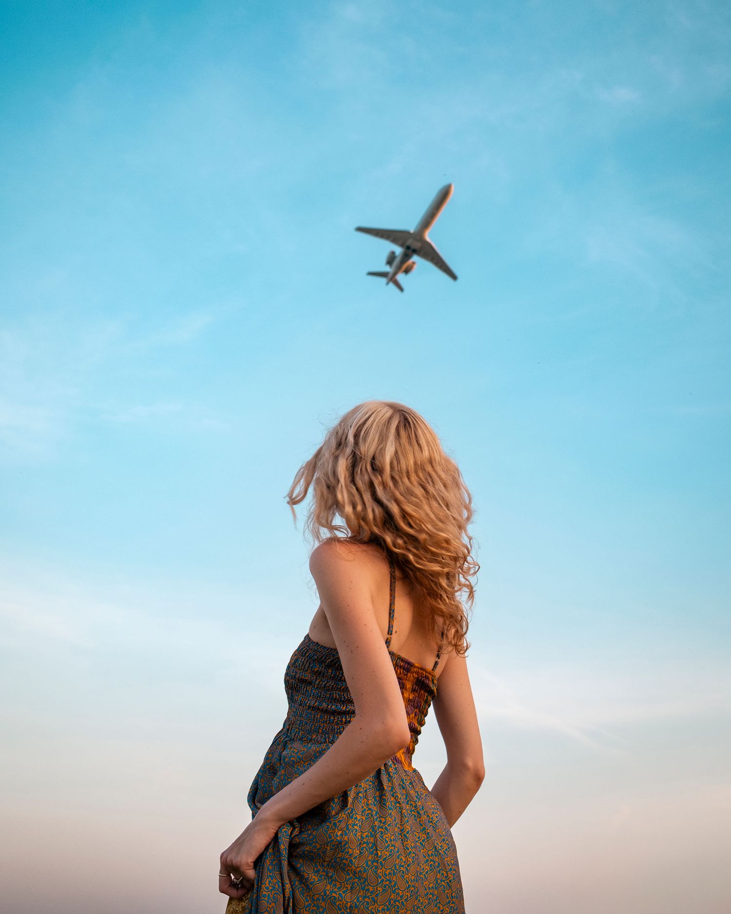 Woman in floral dress with curly hair watching airplane fly overhead against blue sky.