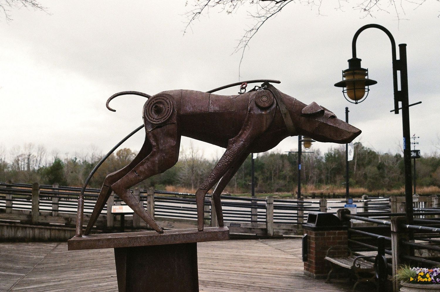 Rustic metal dog sculpture on an outdoor platform with a vintage street lamp against cloudy sky.