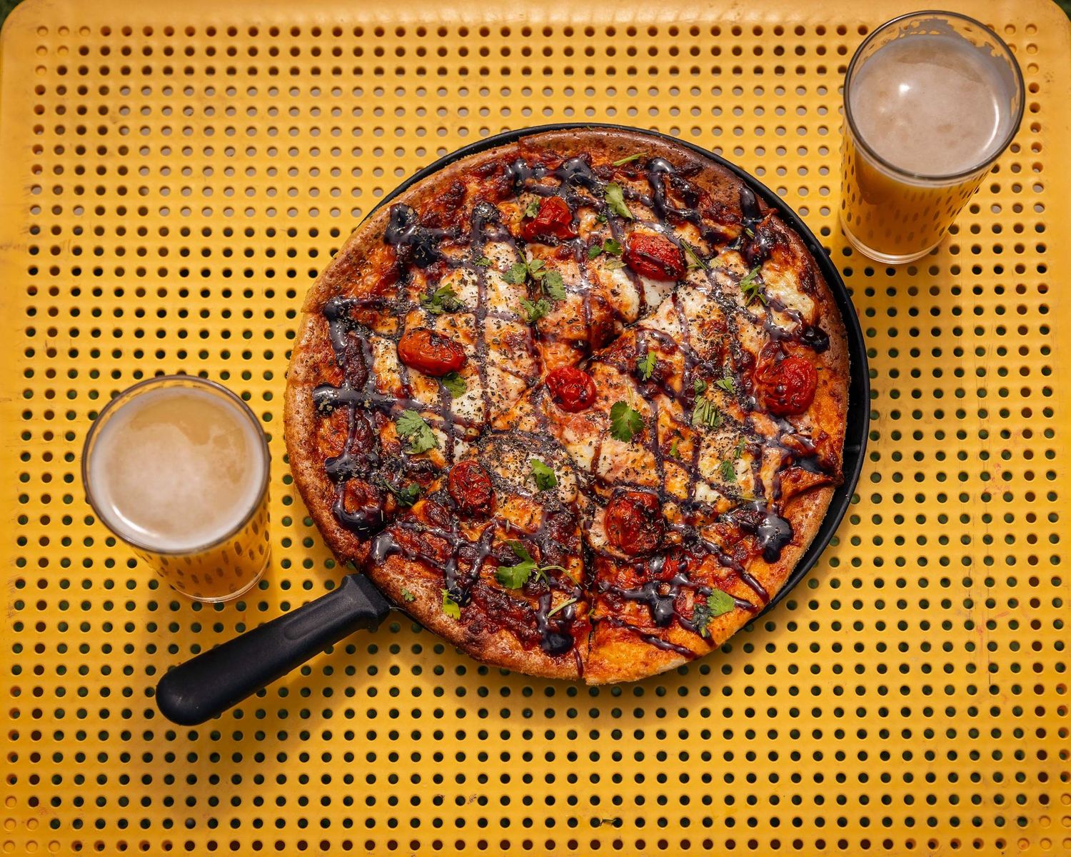 A cast iron skillet pizza topped with tomatoes and herbs served alongside two glasses of beer.
