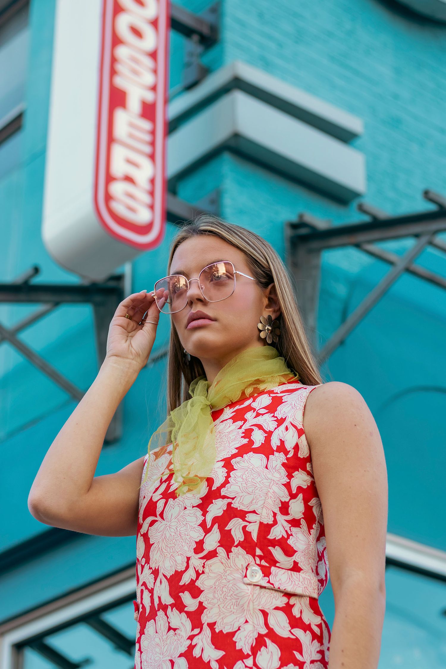 A woman in a red floral dress adjusts pink sunglasses in front of a teal brick building.