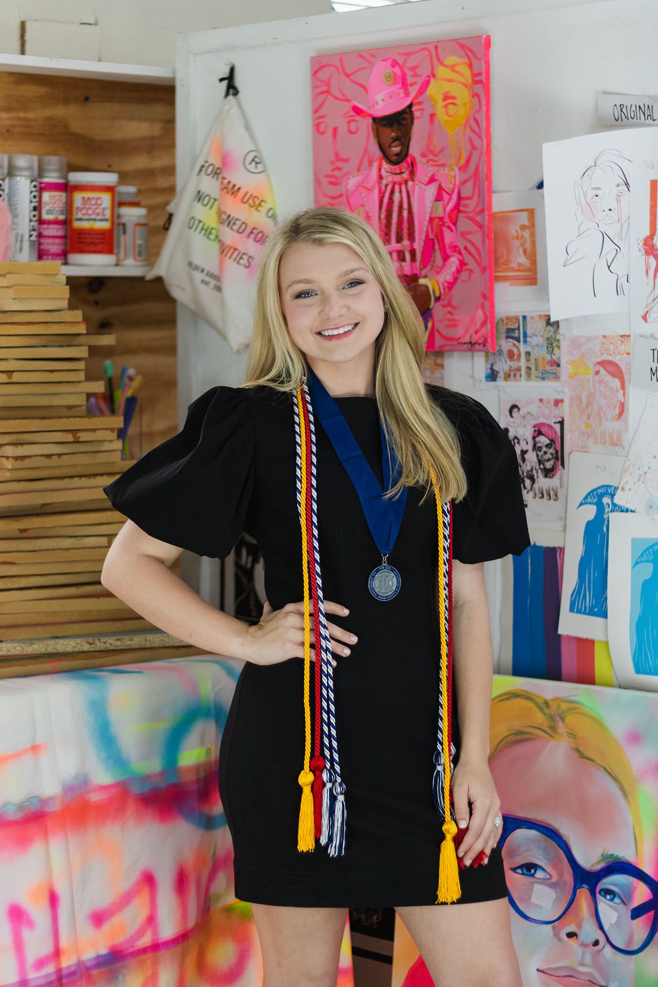 Smiling blonde graduate in black dress displays colorful honor cords and medal in vibrant art studio.