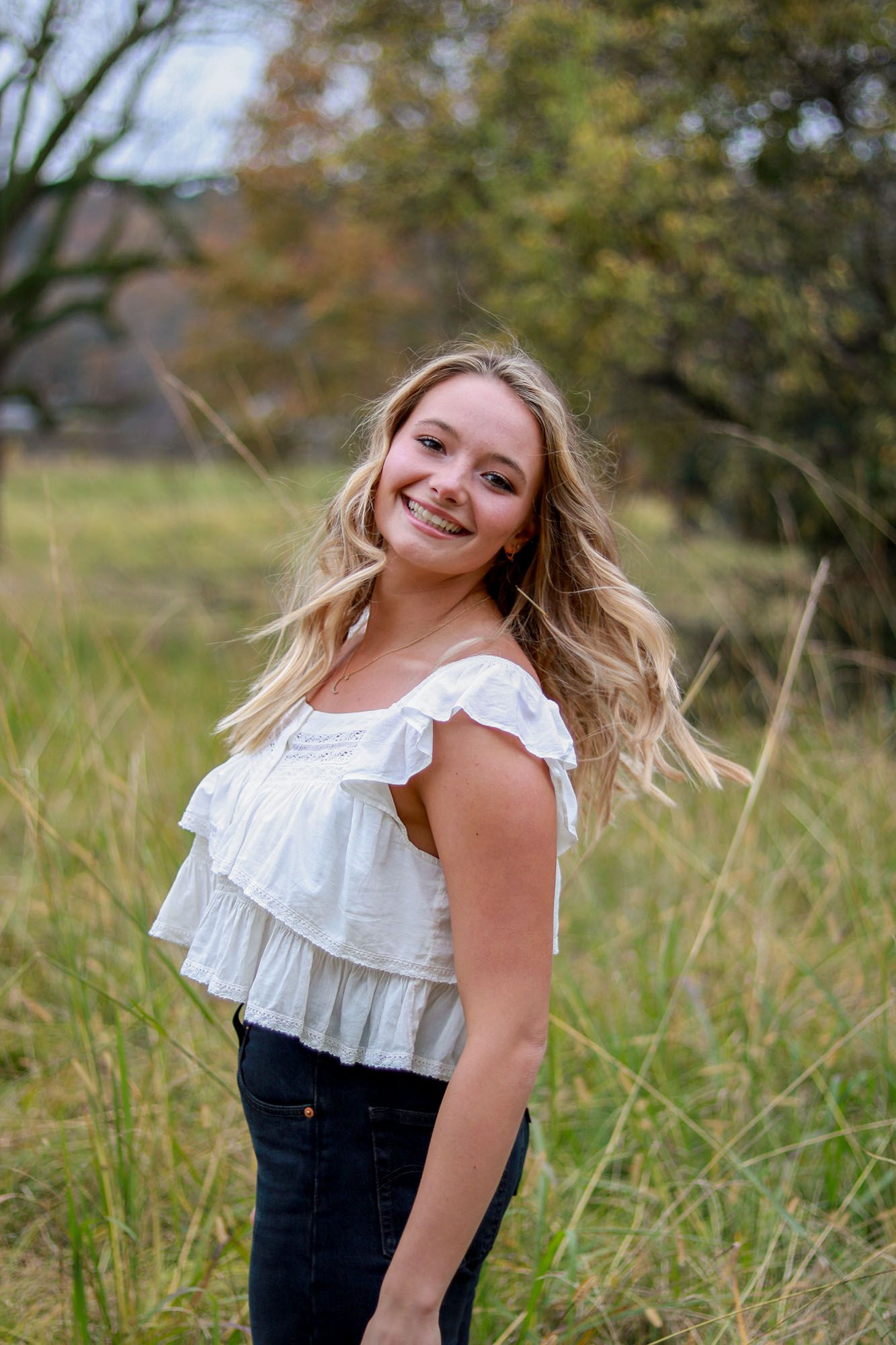 Cheerful blonde teenage girl in white ruffled top posing in tall grass field outdoors.