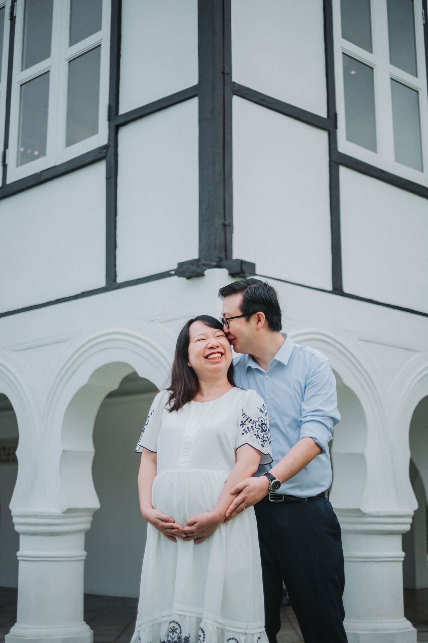 A romantic couple embraces in front of a white colonial building with arched columns and windows.