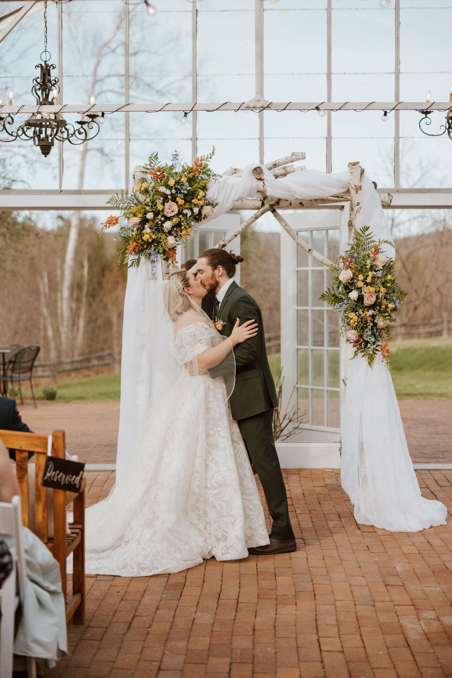 Bride and groom sharing first kiss under a floral birch arch at an outdoor greenhouse ceremony.