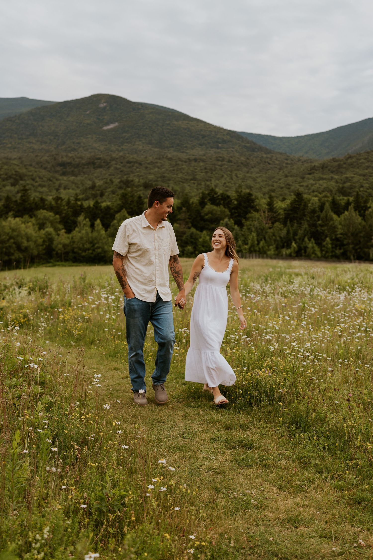Happy couple walking hand in hand through summer wildflower meadow with green mountain scenery.