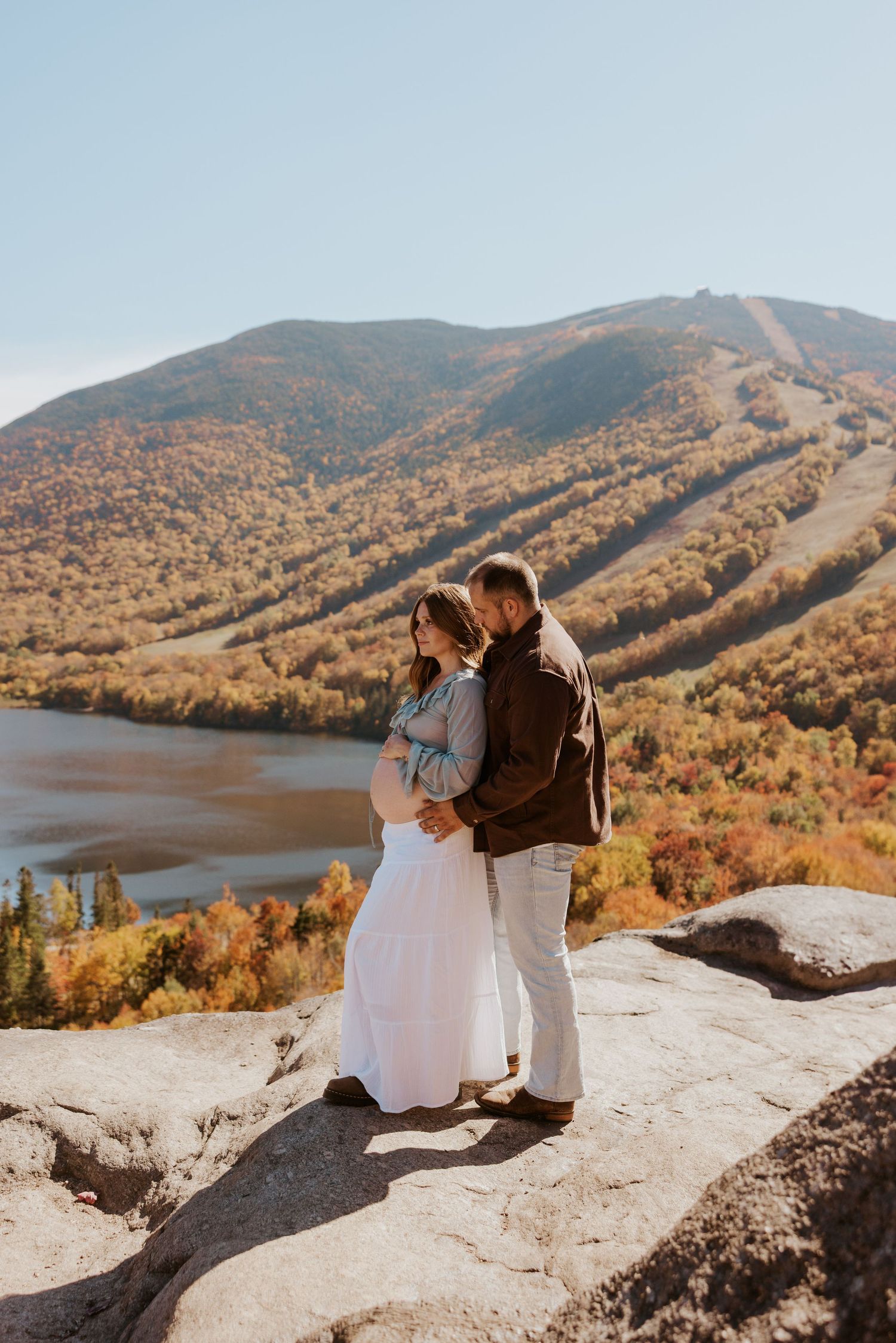 Couple embracing on rocky overlook with colorful autumn mountain and lake landscape behind them.