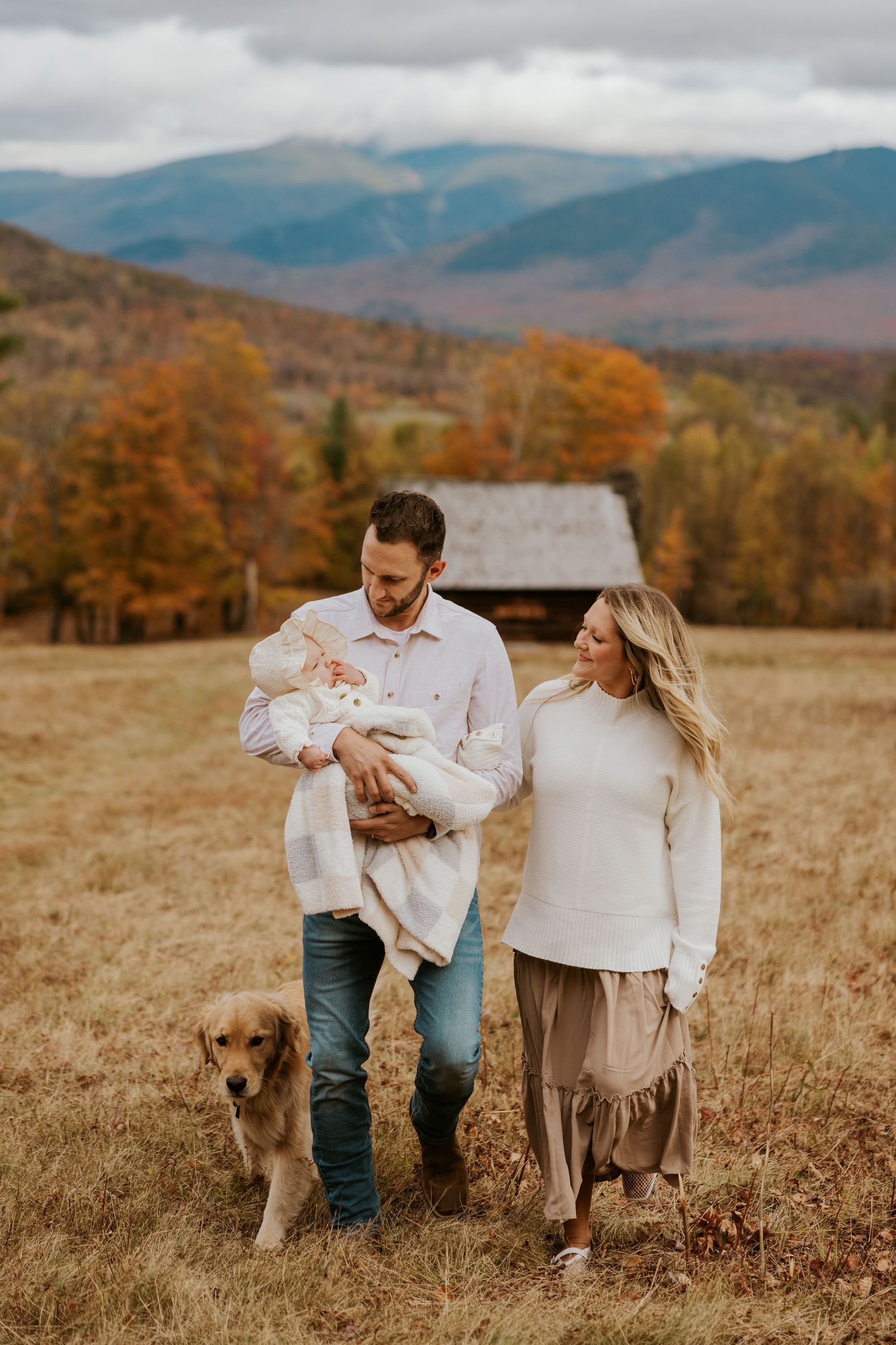 Family with golden retriever walking in autumn mountain field, father holding swaddled newborn.