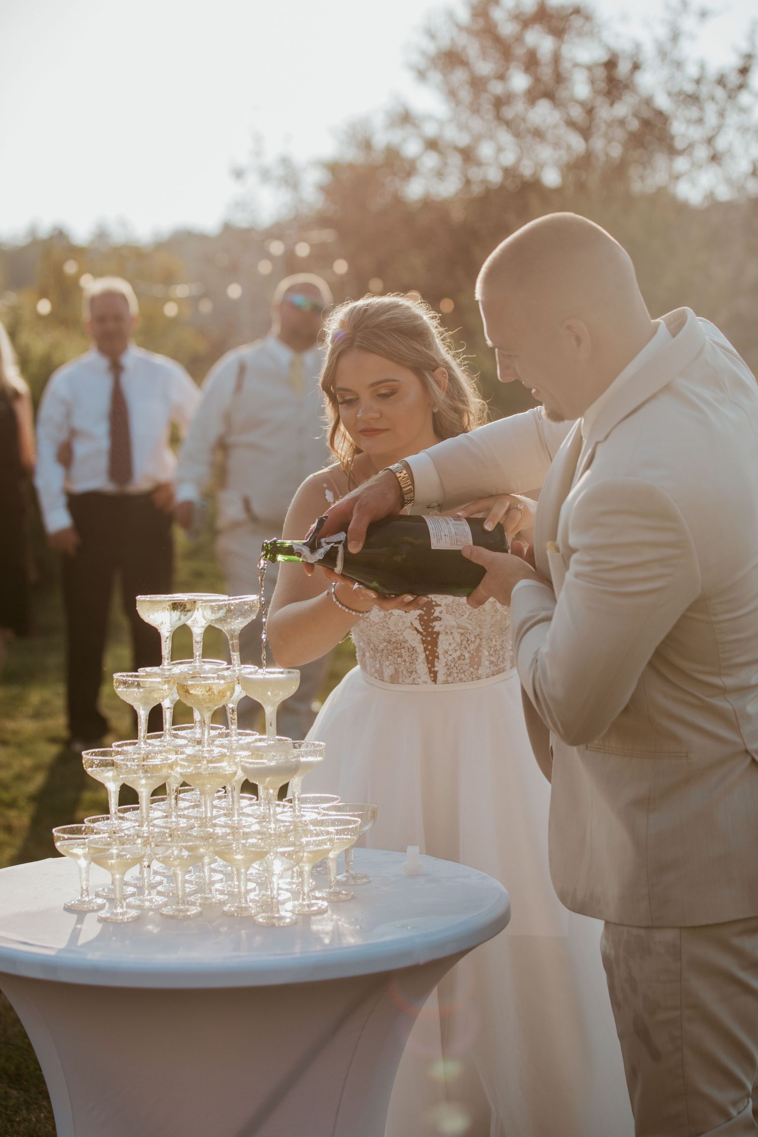 Bride and groom together pouring champagne tower at golden hour outdoor wedding reception.