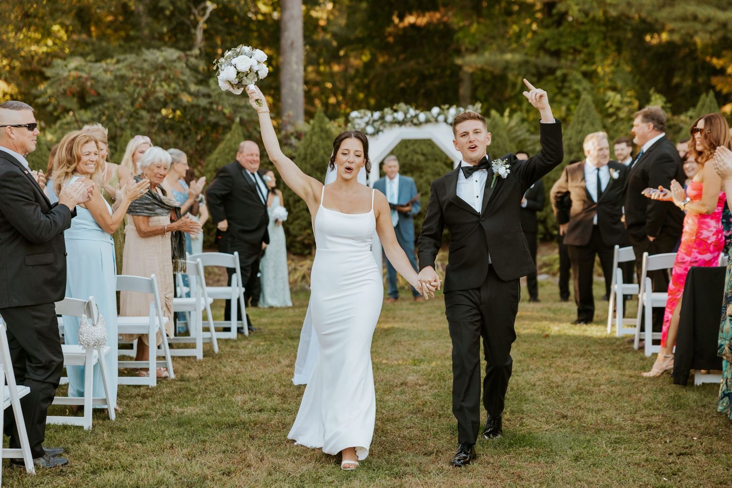 Joyful newlyweds celebrate walking back down aisle with guests applauding at outdoor wedding.