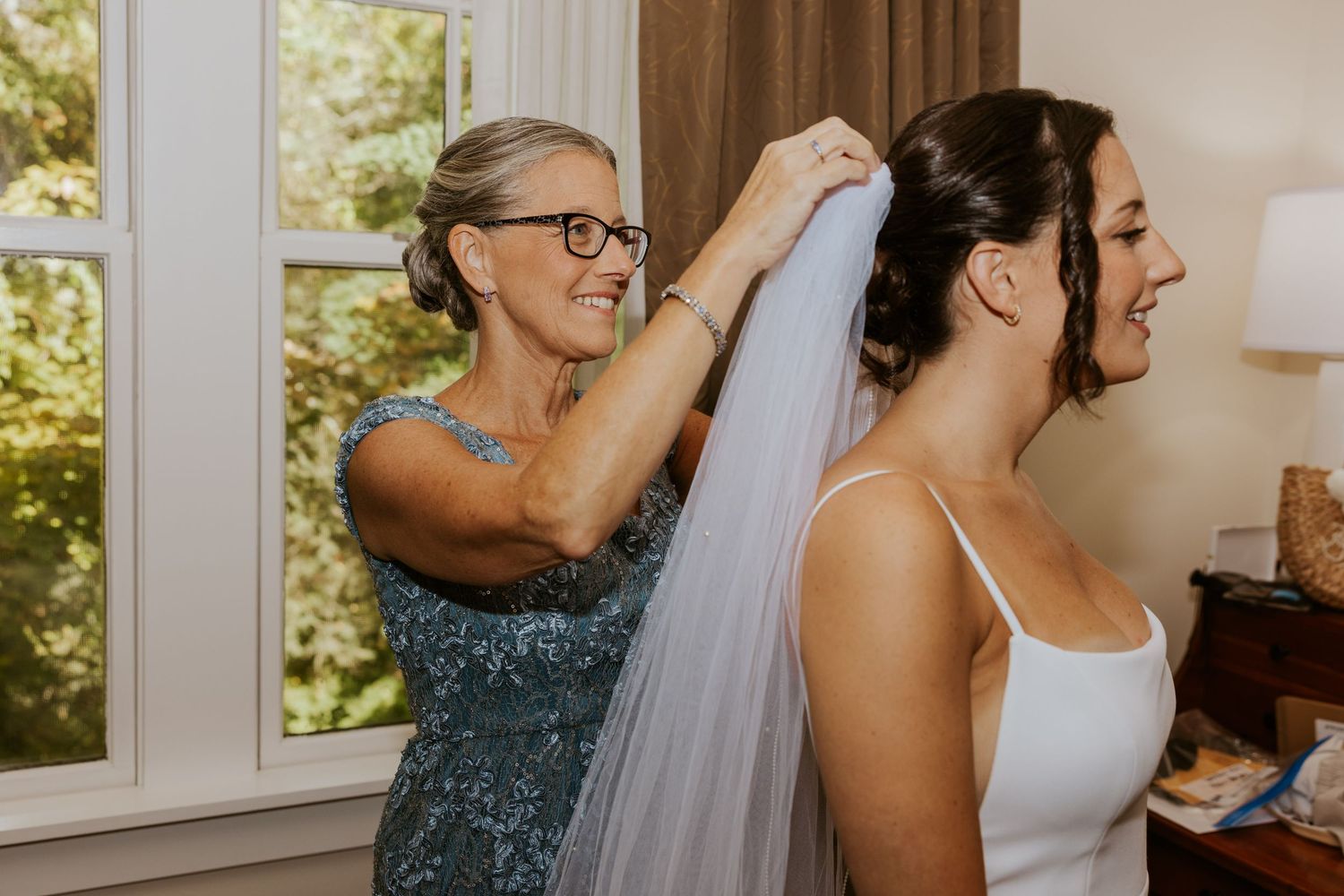 Mother placing veil on brides updo hairstyle while smiling in a warmly lit hotel room.